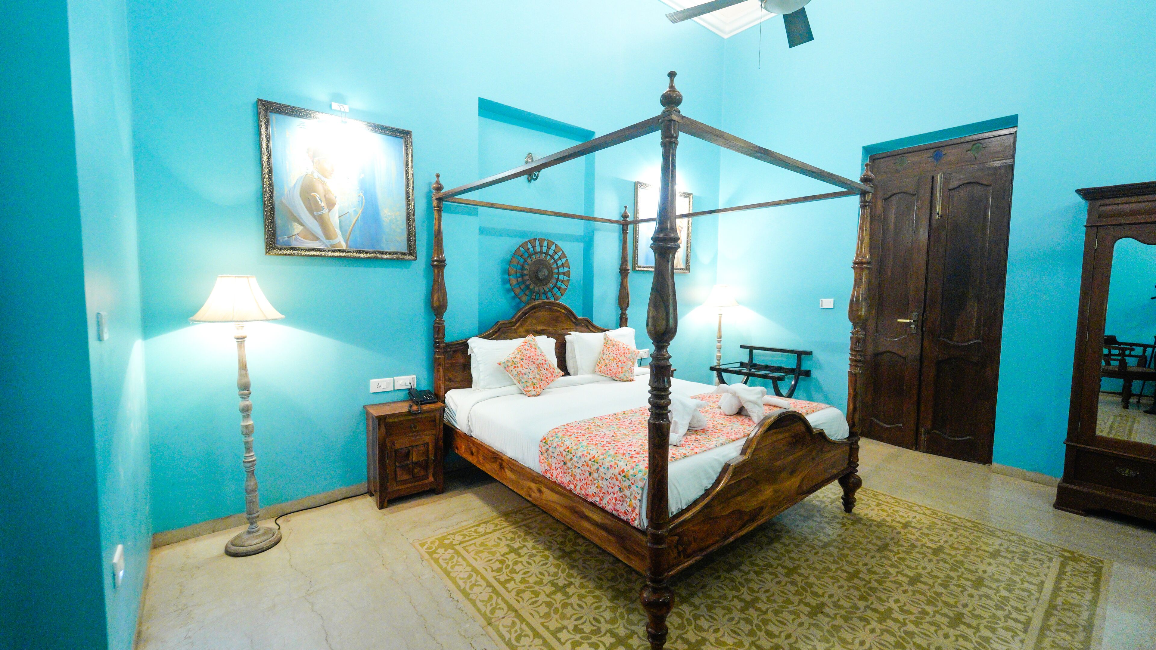 A four-poster bed with a floral bedspread, a bedside table with a lamp, a wall clock, and a mirror.