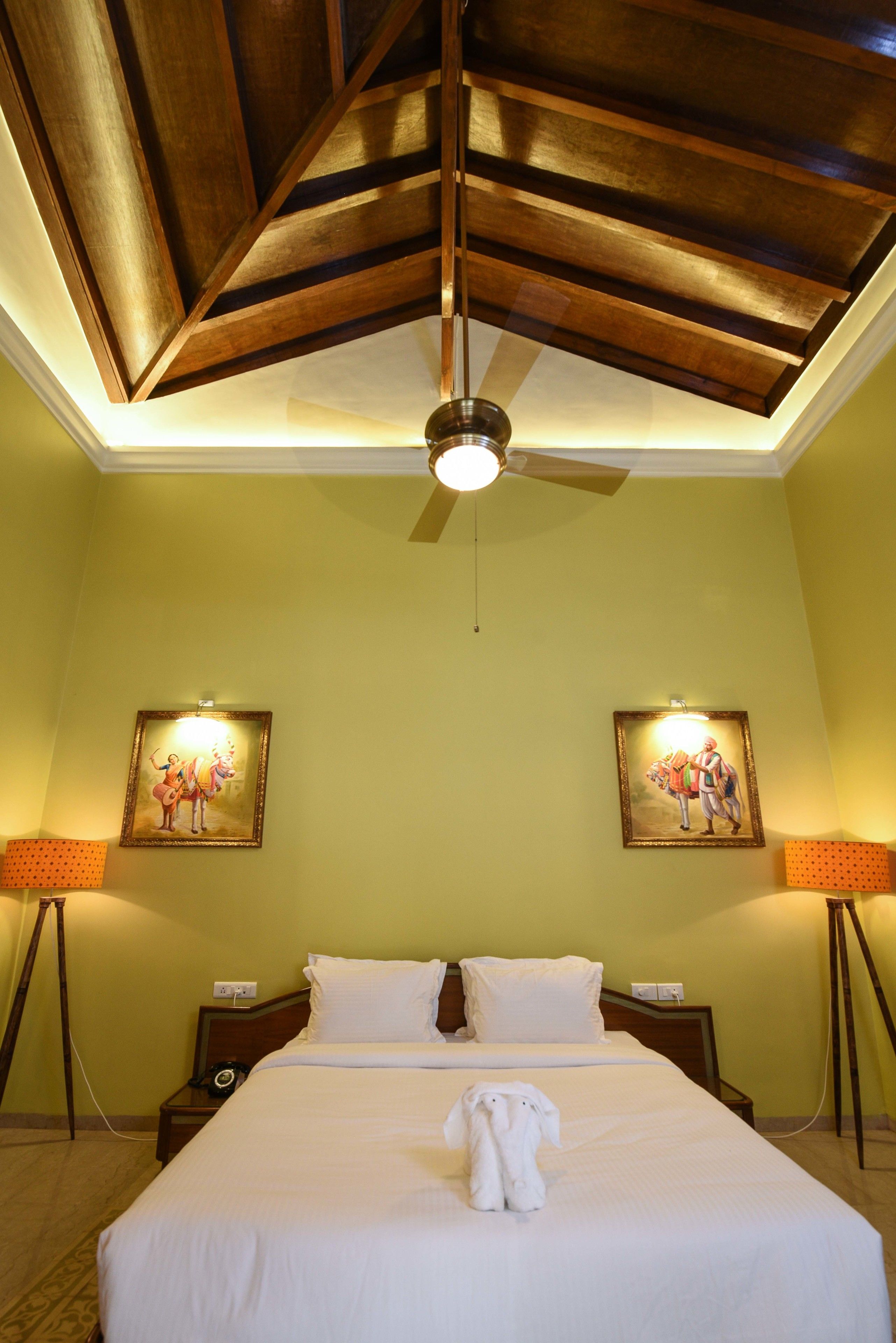 A hotel room with a large bed, two paintings on the walls, a ceiling fan, and a nightstand with a lamp.