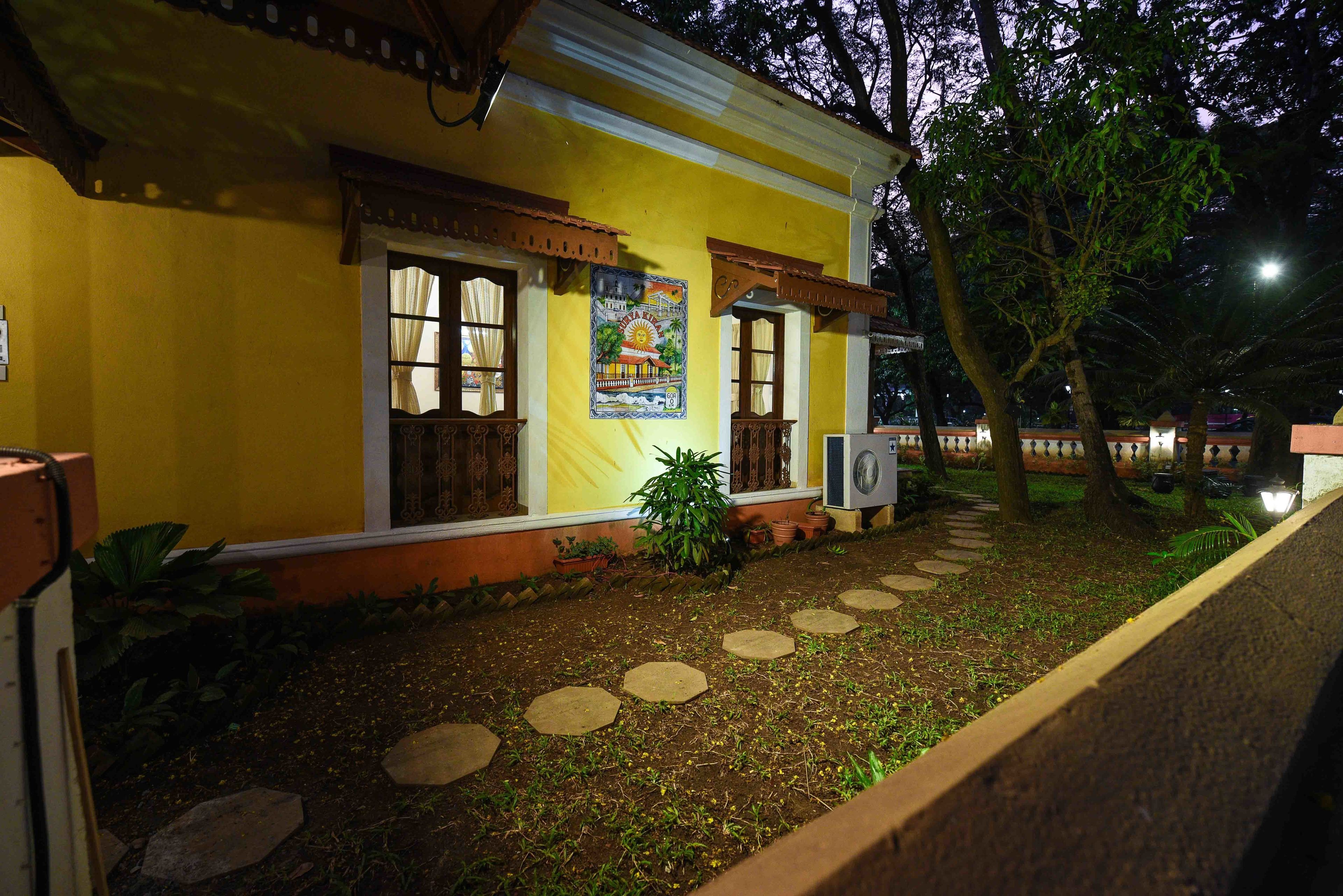 A yellow building with a garden path leading to the entrance, surrounded by trees and plants.