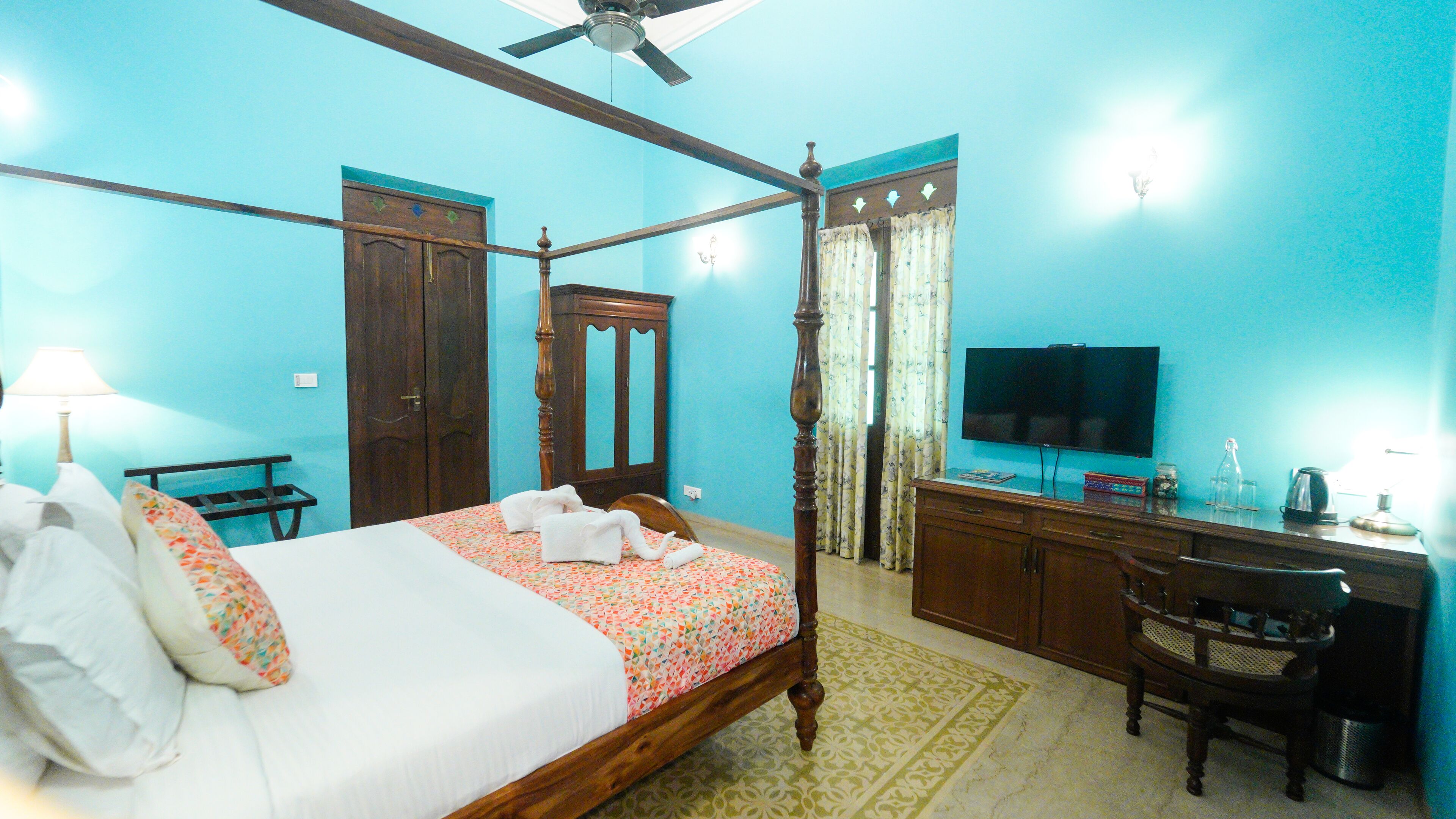 A four-poster bed with a floral bedspread, a wooden desk with a television, and a desk lamp.