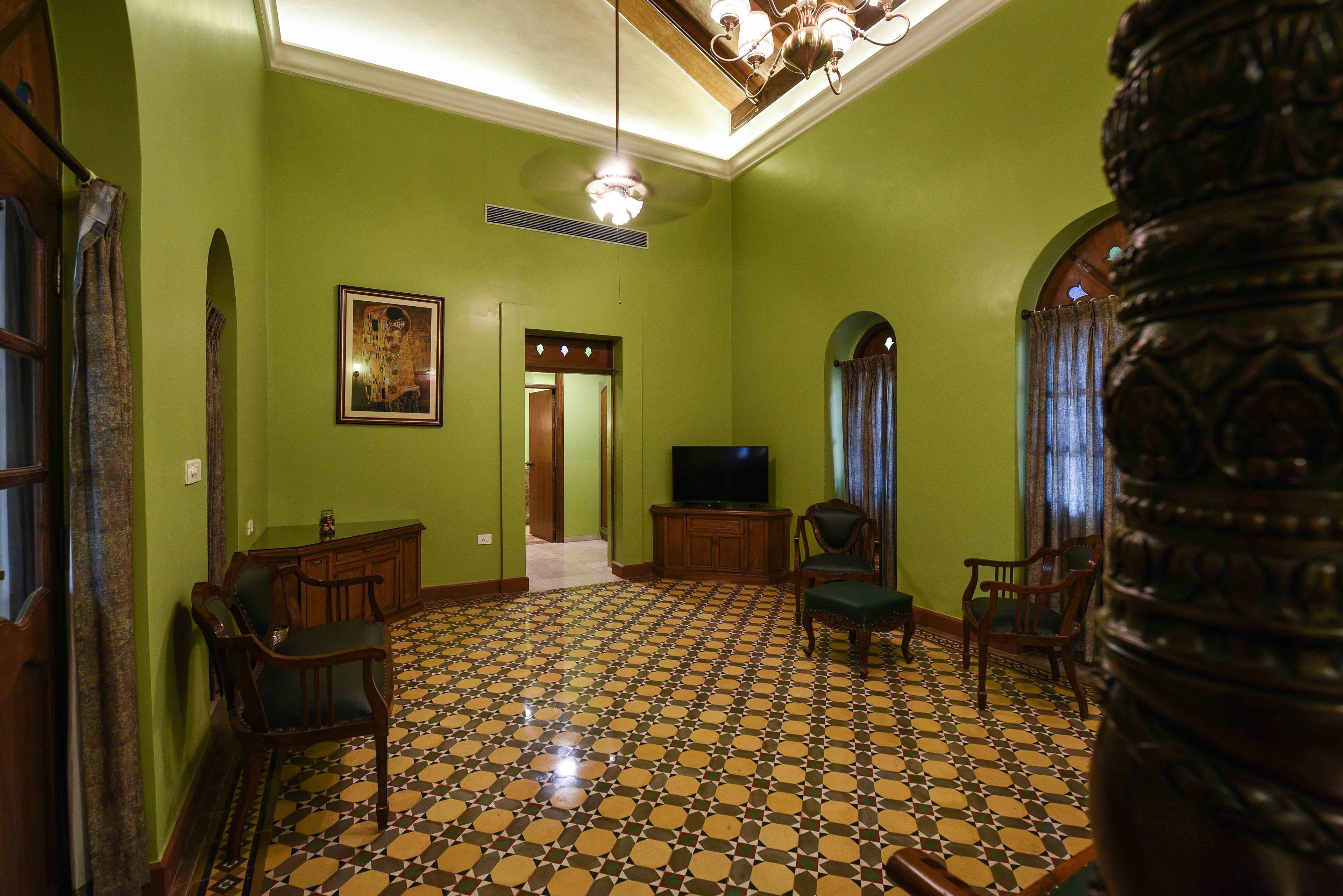 A room with green walls, a patterned floor, and a chandelier. There is a television, a wooden cabinet, and two chairs.