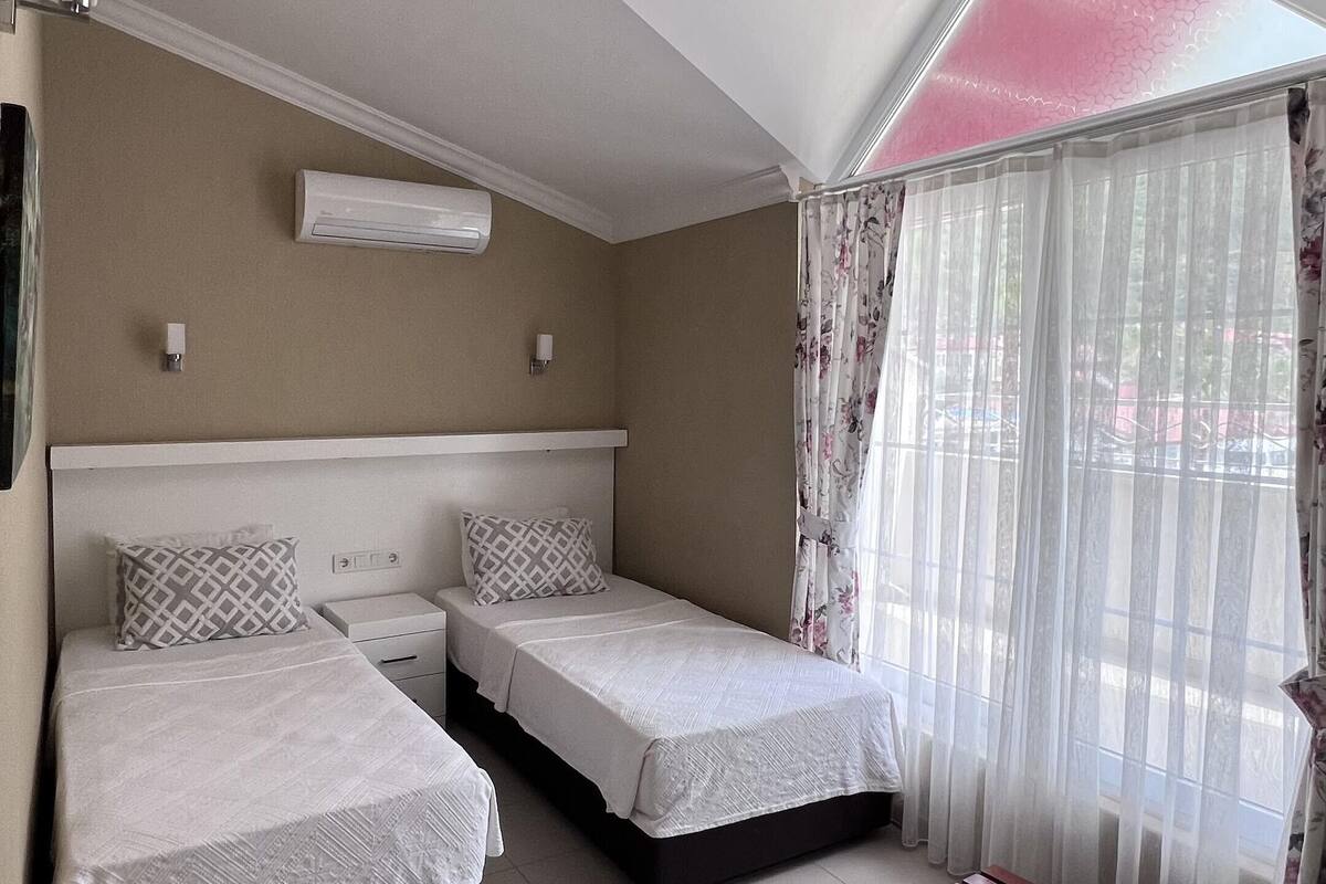 A bedroom with two beds, a bench, a ceiling fan, and a window with sheer curtains.