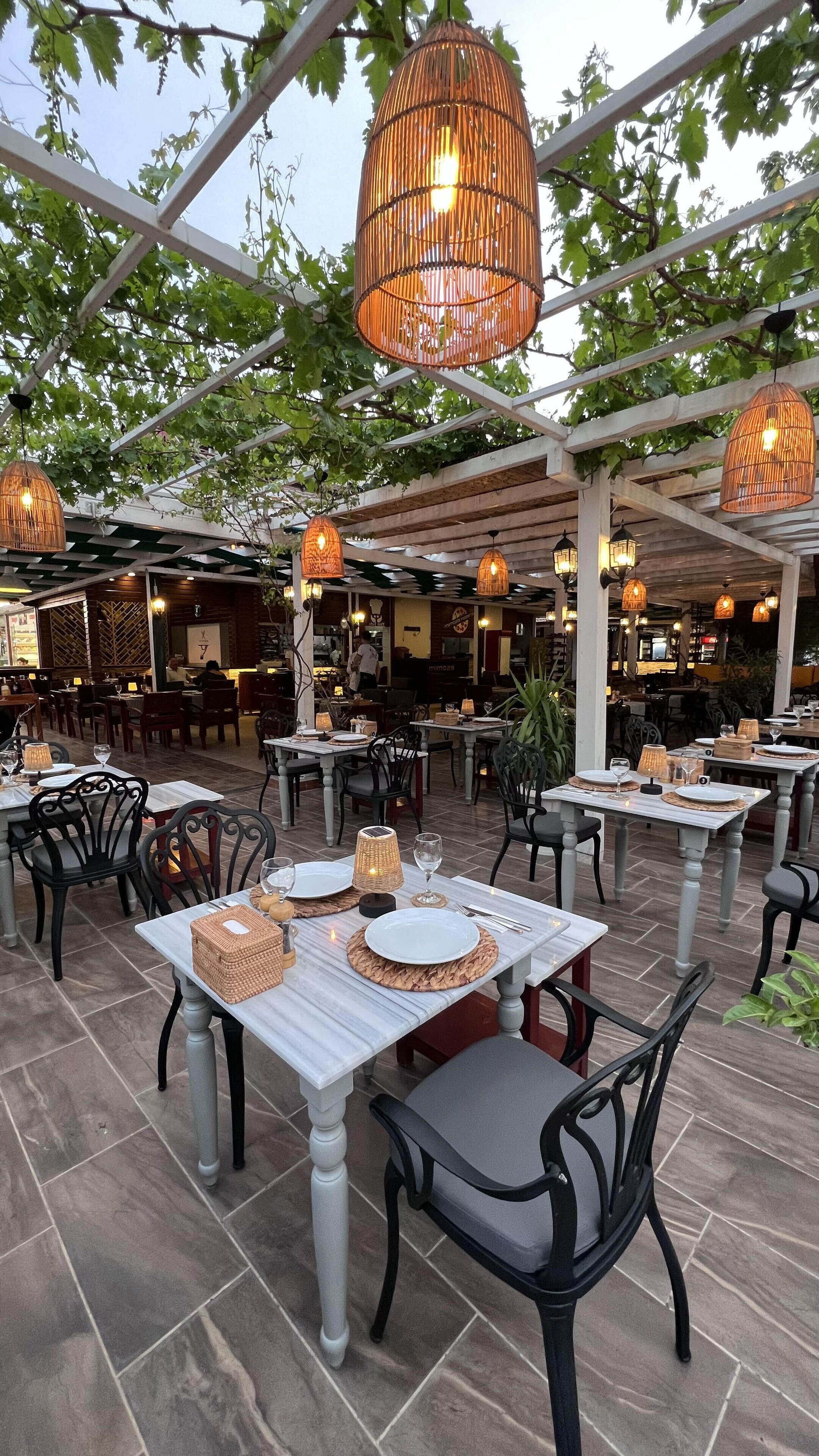 An outdoor restaurant with tables set for dining, surrounded by greenery and hanging wicker lamps.
