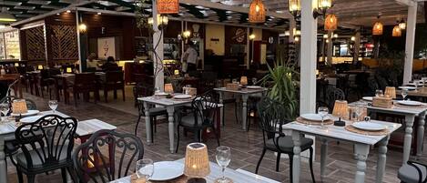 Un restaurante al aire libre con mesas dispuestas para comer, rodeado de follaje y lámparas de mimbre colgantes.