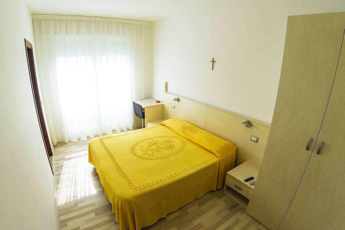 Una habitación de hotel con cama, mesita de noche, armario y ventana con cortinas.