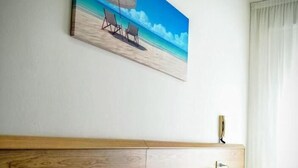 A hotel room with a wooden headboard, a bed with white linens, a painting of a beach scene, and a nightstand with a lamp and decorative items. - Hotel Souvenir (Caorle)