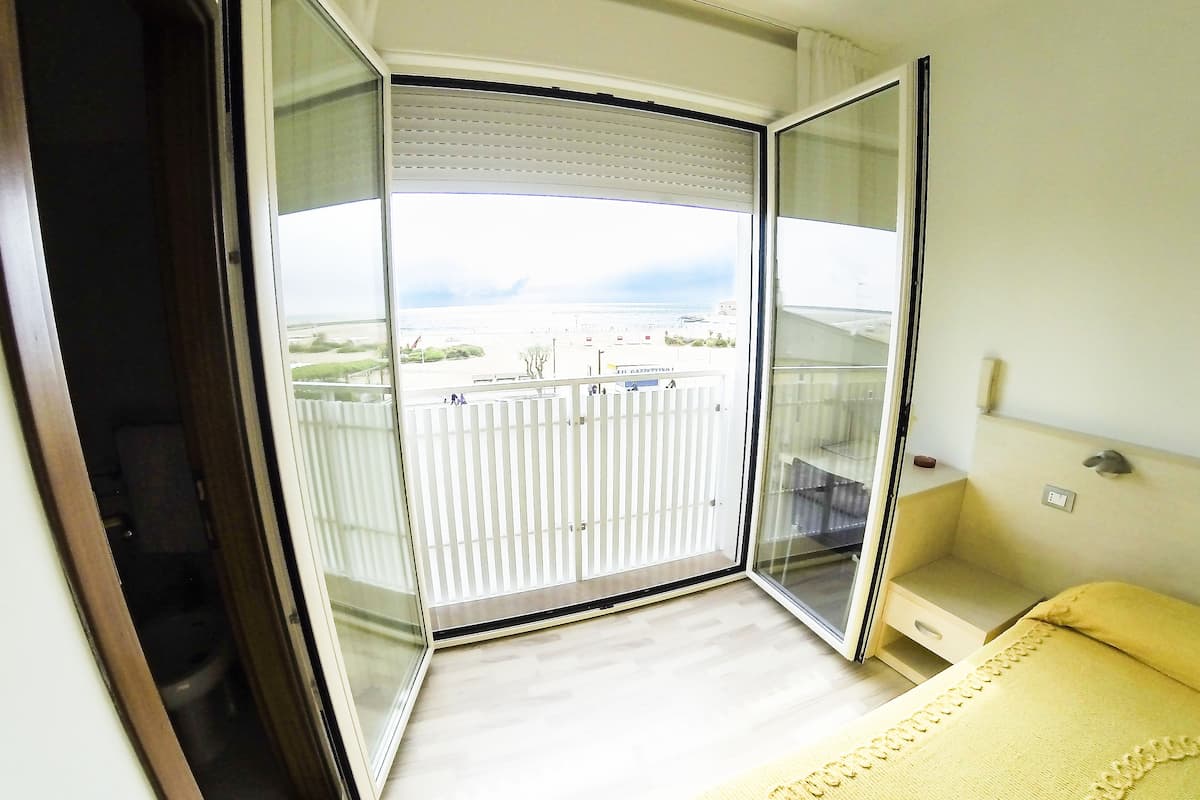 Una habitación con cama, una mesita de noche pequeña, una puerta de vidrio que da a un balcón exterior y vistas al mar.