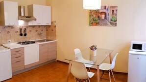 A compact kitchen with a gas stove, white cabinets, a microwave, and a dining area with a glass table and white chairs. - Mosaico Aparthotel (Modena)