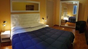 A bedroom with a large bed, a nightstand with a lamp, a mirror, and a door leading to another room. - Mosaico Aparthotel (Modena)