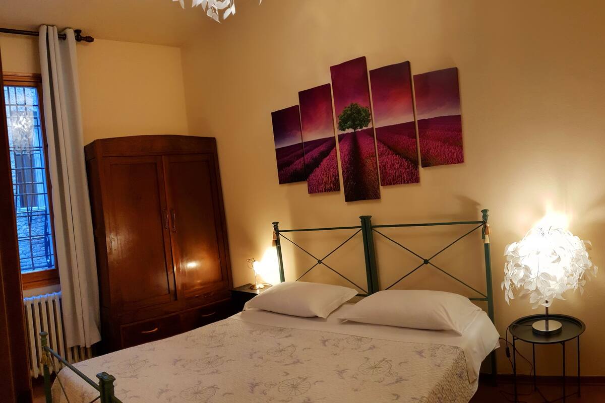 Une chambre à coucher comprenant un lit, une armoire en bois, un radiateur, une fenêtre avec des rideaux et un mur orné d’un tableau représentant un paysage.
