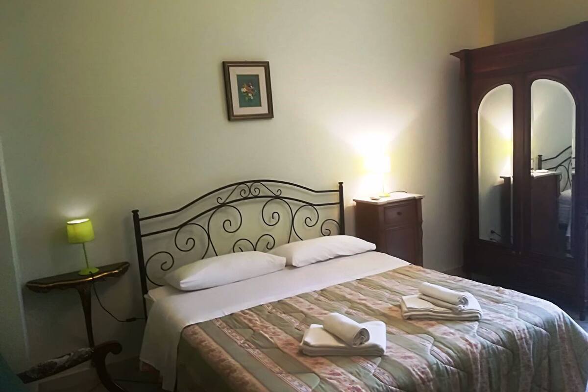 Una cama bien hecha con cubrecama estampado y toallas enrolladas, una lámpara de noche, un cuadro enmarcado en la pared y un mueble espejo.