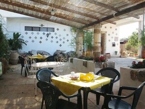 Une terrasse extérieure avec une table et des chaises, une pergola et, au fond, un bâtiment blanc orné de carreaux décoratifs.