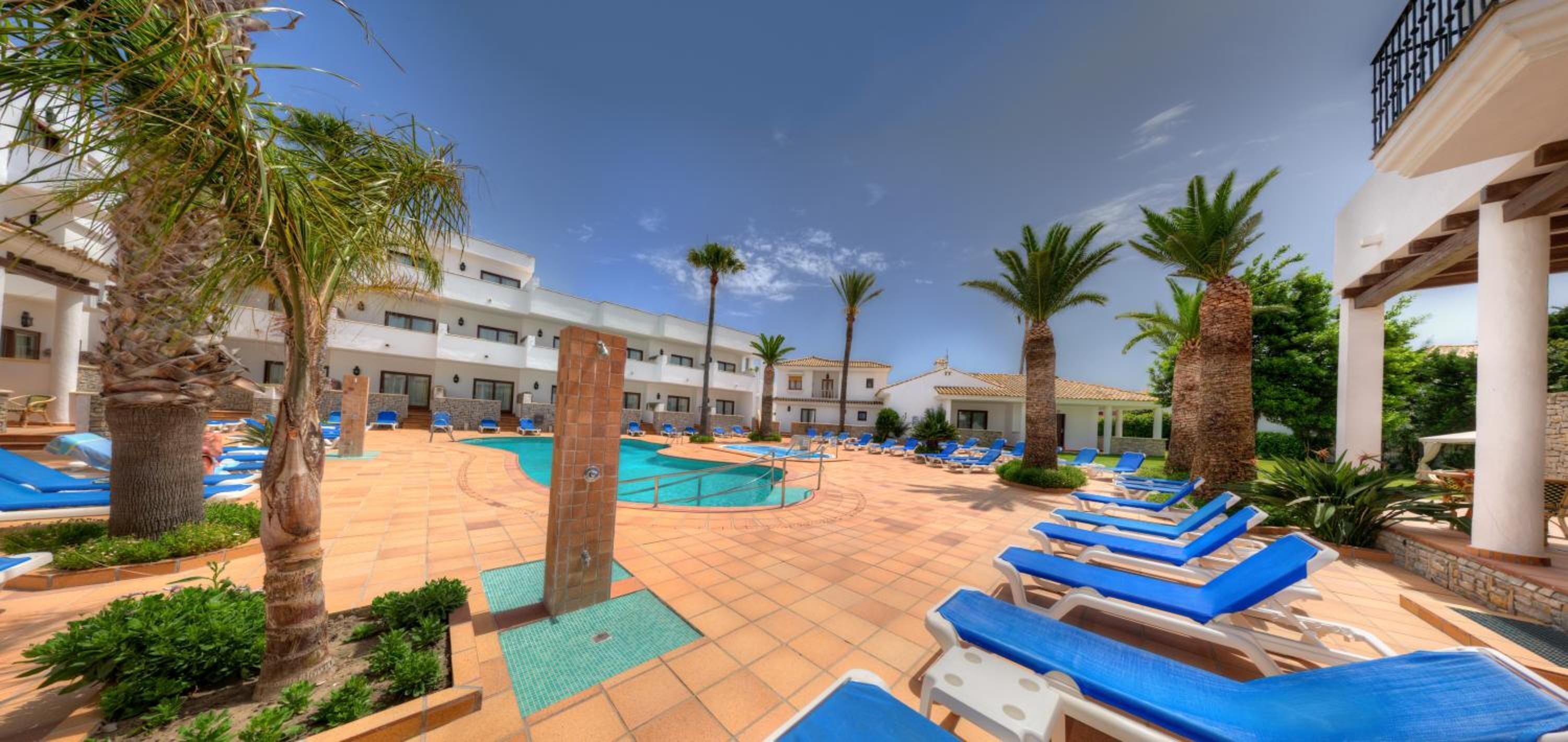 Un espace piscine dans un complexe hôtelier comprenant plusieurs piscines, des chaises longues, des palmiers et une terrasse carrelée.