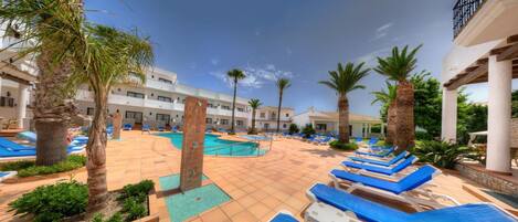 Un espace piscine dans un complexe hôtelier comprenant plusieurs piscines, des chaises longues, des palmiers et une terrasse carrelée.