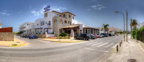 Un hotel con fachada blanca, varios balcones y un estacionamiento con varios coches.
