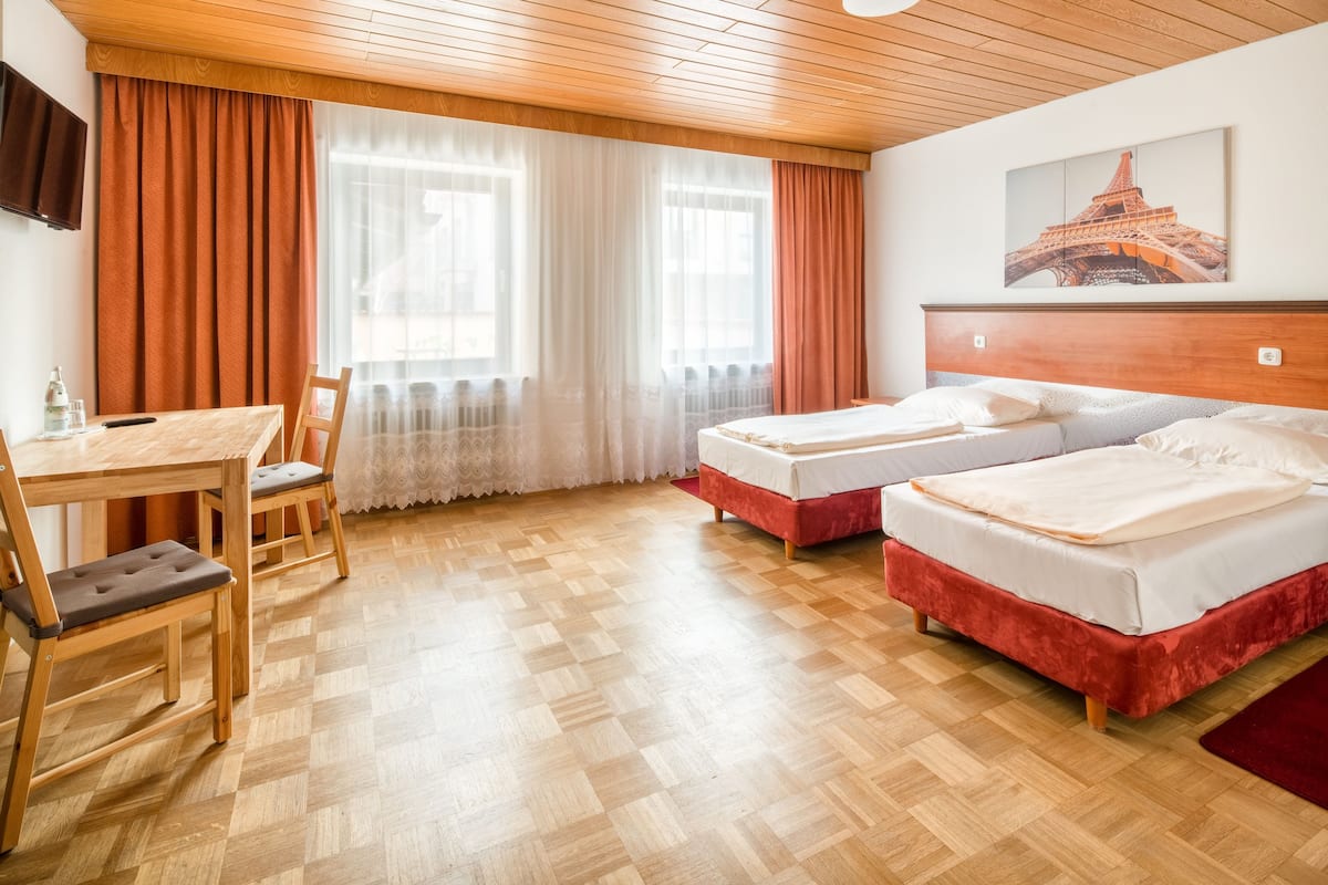 Une chambre d’hôtel avec un plancher en bois, deux lits avec une literie rouge, un bureau en bois avec une chaise, un téléviseur fixé au mur et une fenêtre avec des rideaux.