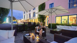 Un espace extérieur aménagé avec des meubles en osier et des parasols, une femme assise sur une chaise, et un bâtiment dont les fenêtres sont illuminées en arrière-plan.