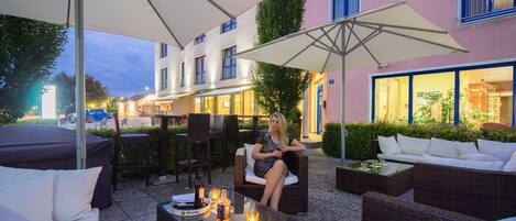 An outdoor seating area with wicker furniture and umbrellas, a woman sitting on a chair, and a building with illuminated windows in the background.
