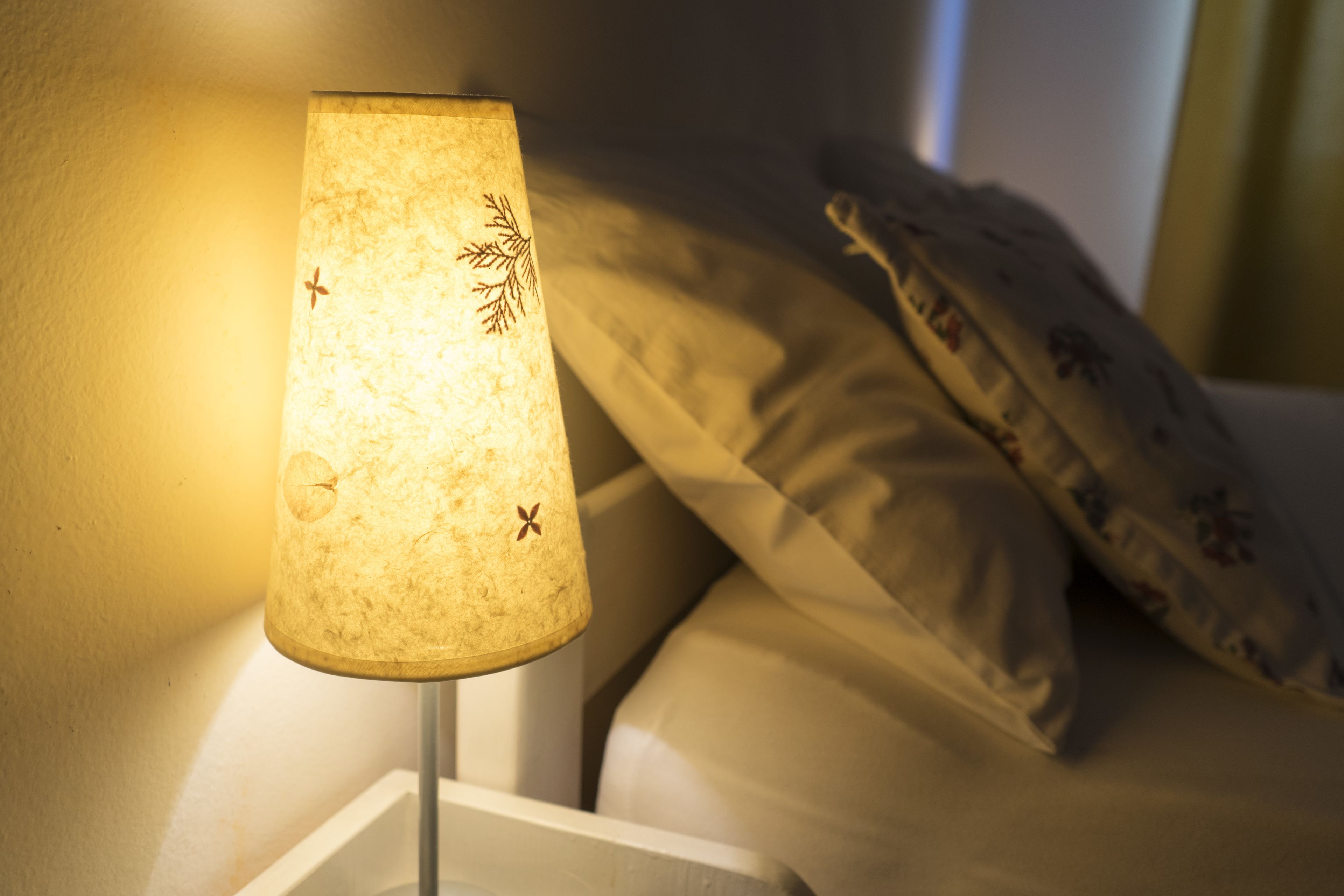 A bedside lamp with a textured lampshade, a white bedside table, and a bed with a floral pillow.