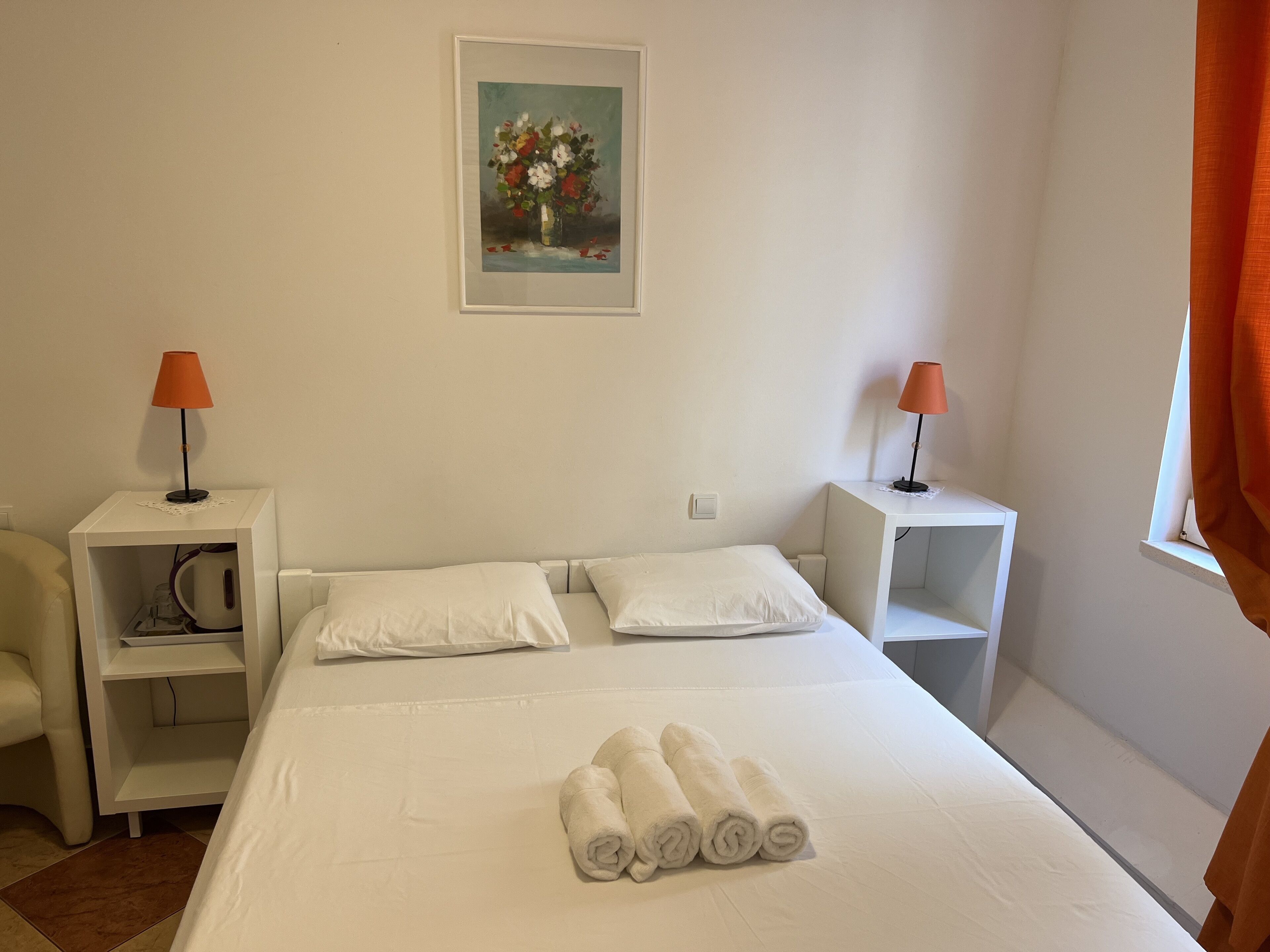 Una cama bien hecha con sábanas blancas y toallas enrolladas, dos mesitas de noche con lámparas, un aparador blanco, un cuadro en la pared y una ventana con cortinas.