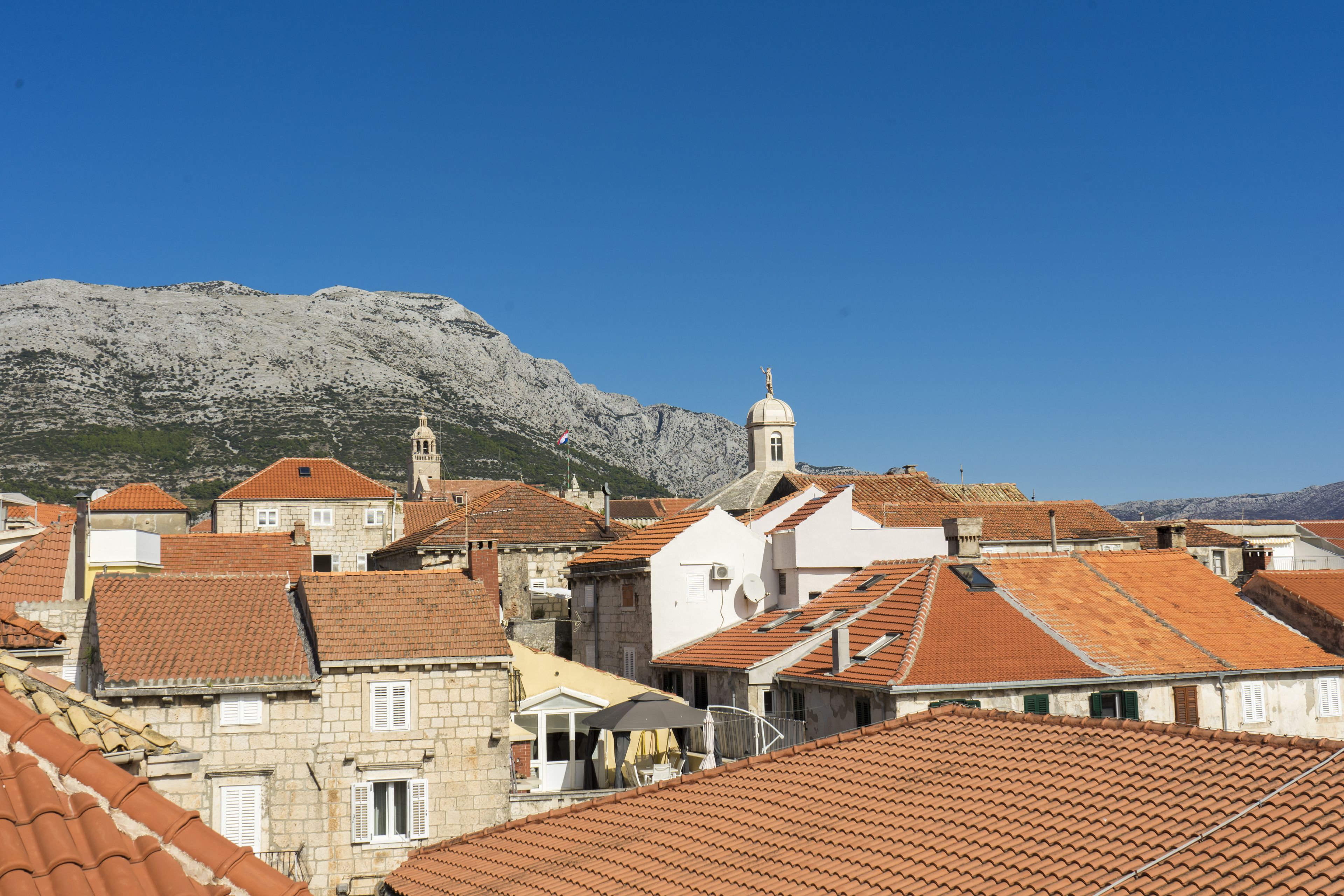 A coastal town with terracotta-roofed buildings, a prominent church with a bell tower, and a mountainous backdrop.