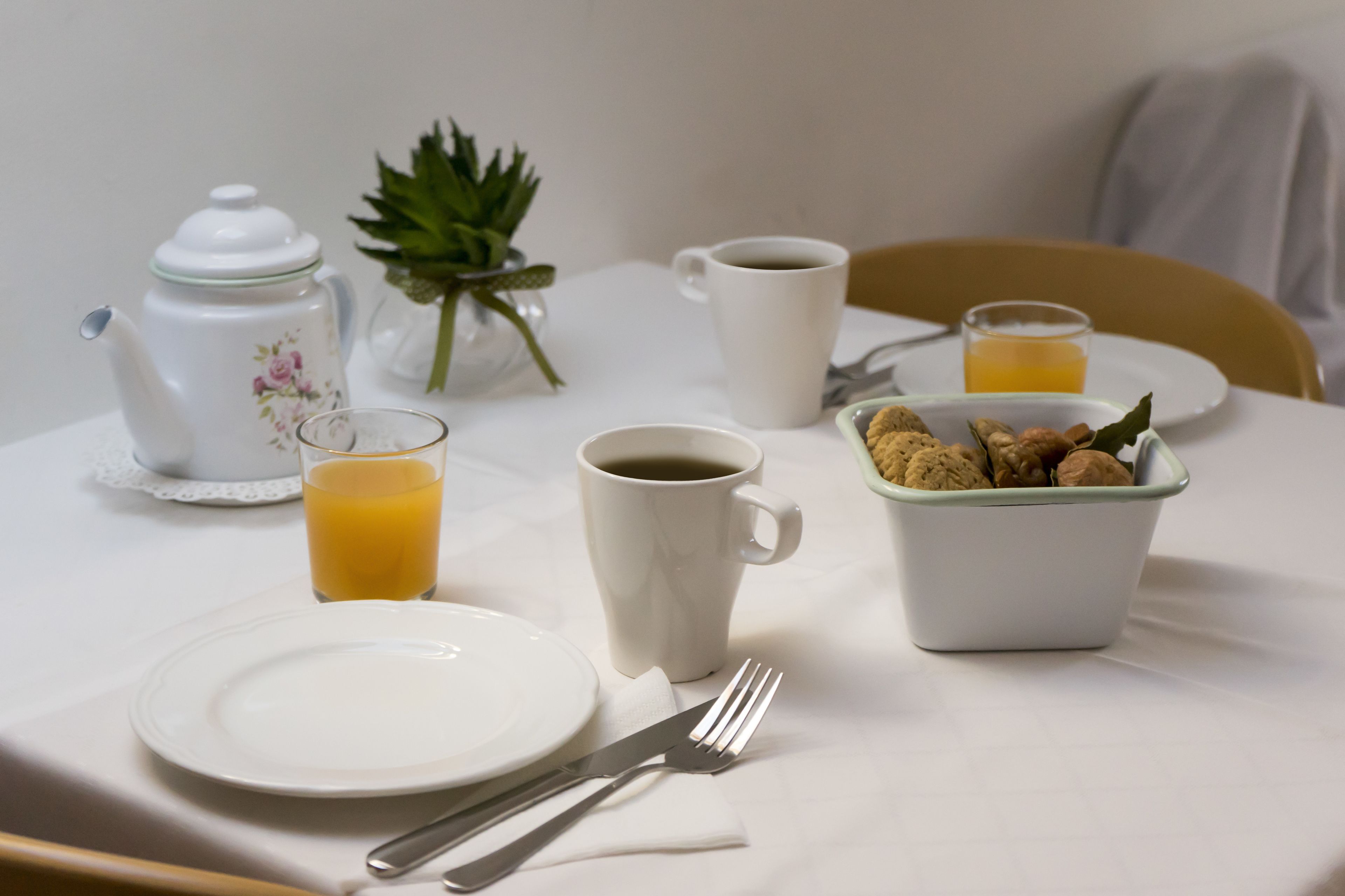 Een ontbijttafel met een theepot, koffiekop, glas jus d'orange, een schaal koekjes en een bord.