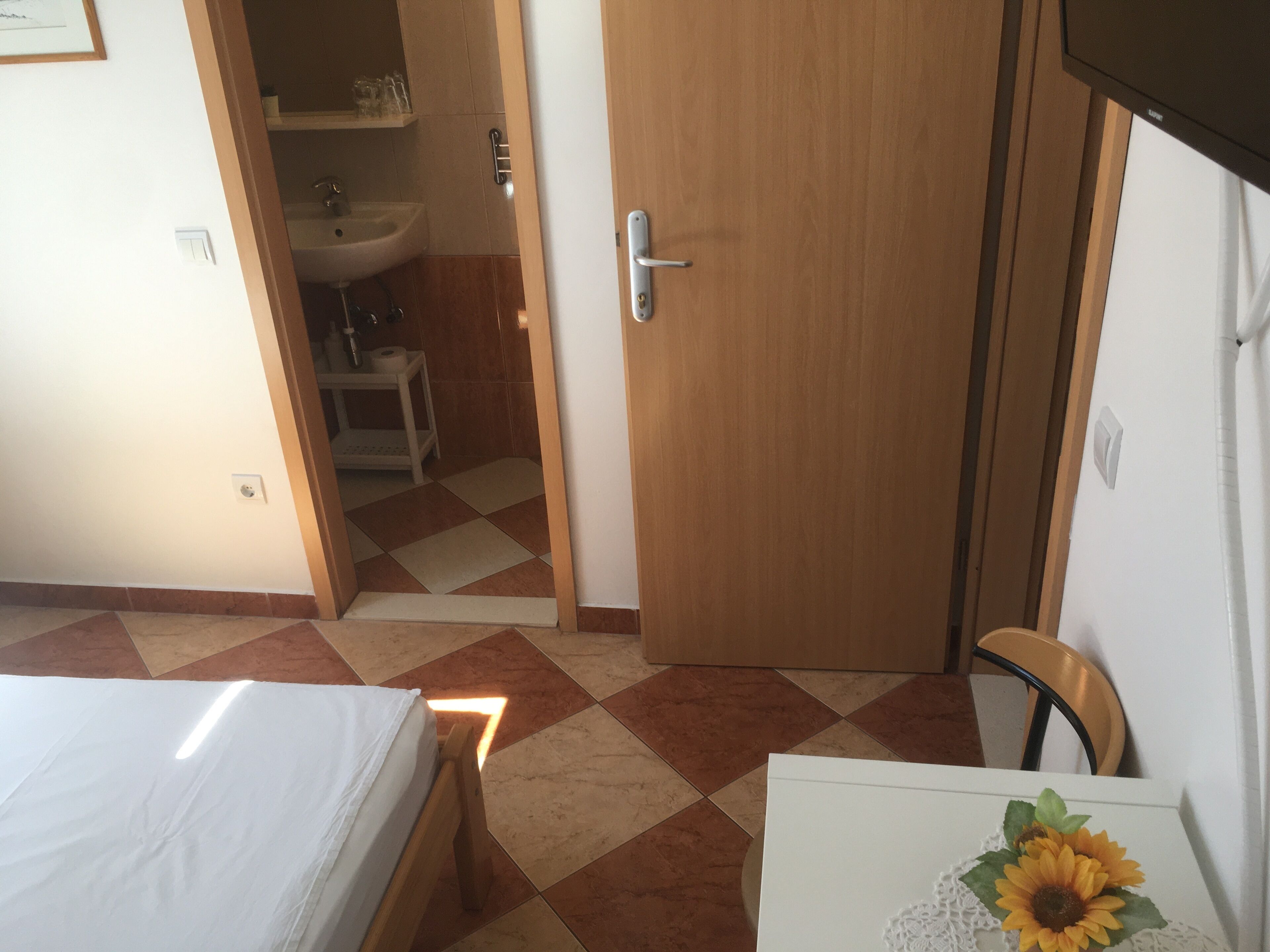 Une chambre d’hôtel avec un lit, un bureau, une chaise, une petite table ornée d’un vase de tournesols, une porte en bois et une salle de bain visible à travers une porte ouverte.