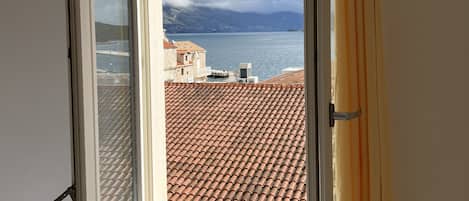 A window with a view of a coastal landscape, a tiled roof, and a curtain.