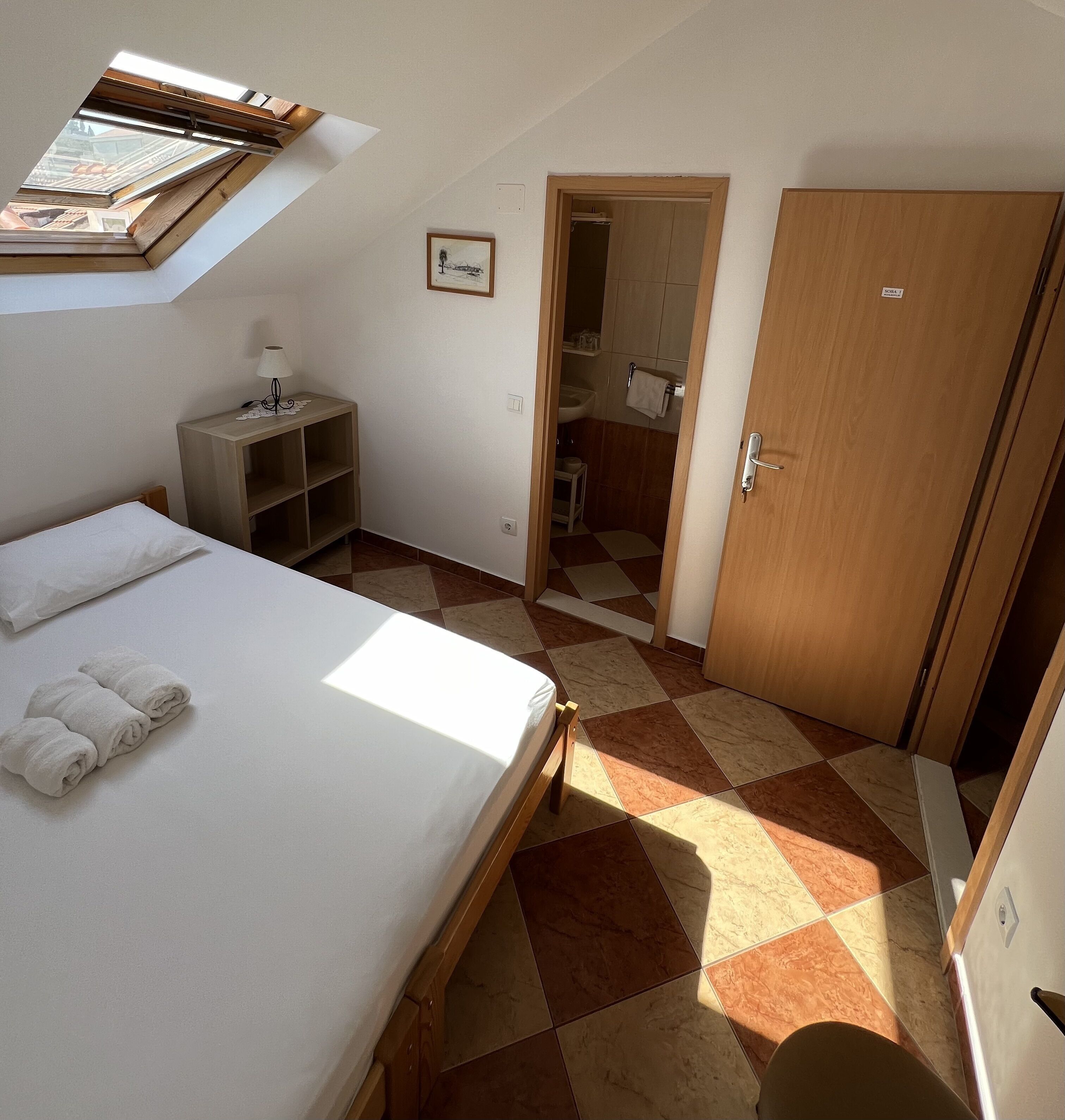 A bedroom with a skylight, a bed with towels, a bedside table, a wardrobe, and a bathroom visible through an open door.