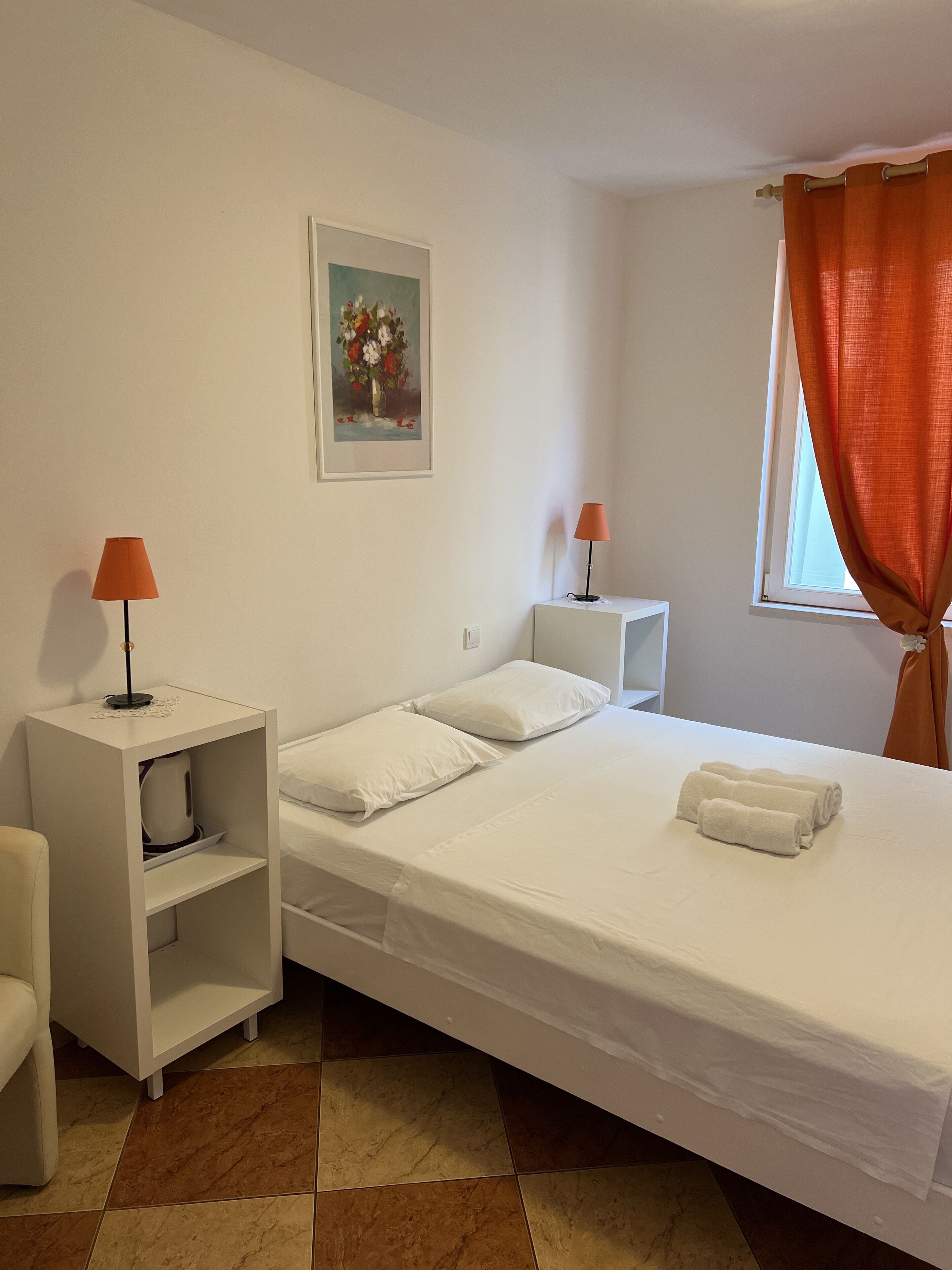 Ein ordentlich eingerichtetes Hotelzimmer mit einem Einzelbett, zwei Nachttischen, einem Sessel, einem Bild an der Wand und einem Fenster mit orangenen Vorhängen.