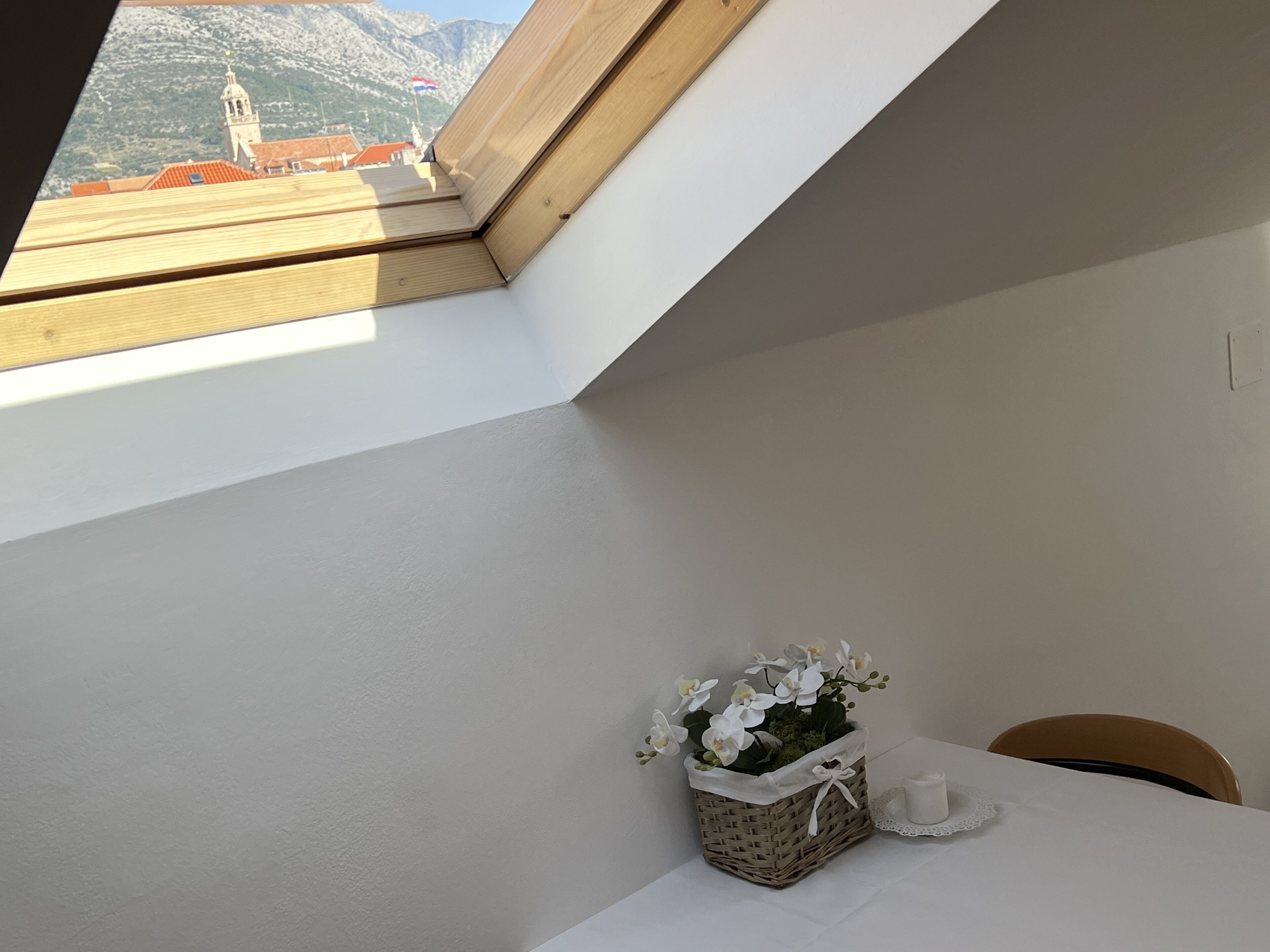 Una habitación con tragaluz, una pequeña mesa con mantel blanco, una cesta de mimbre con flores y una vela.