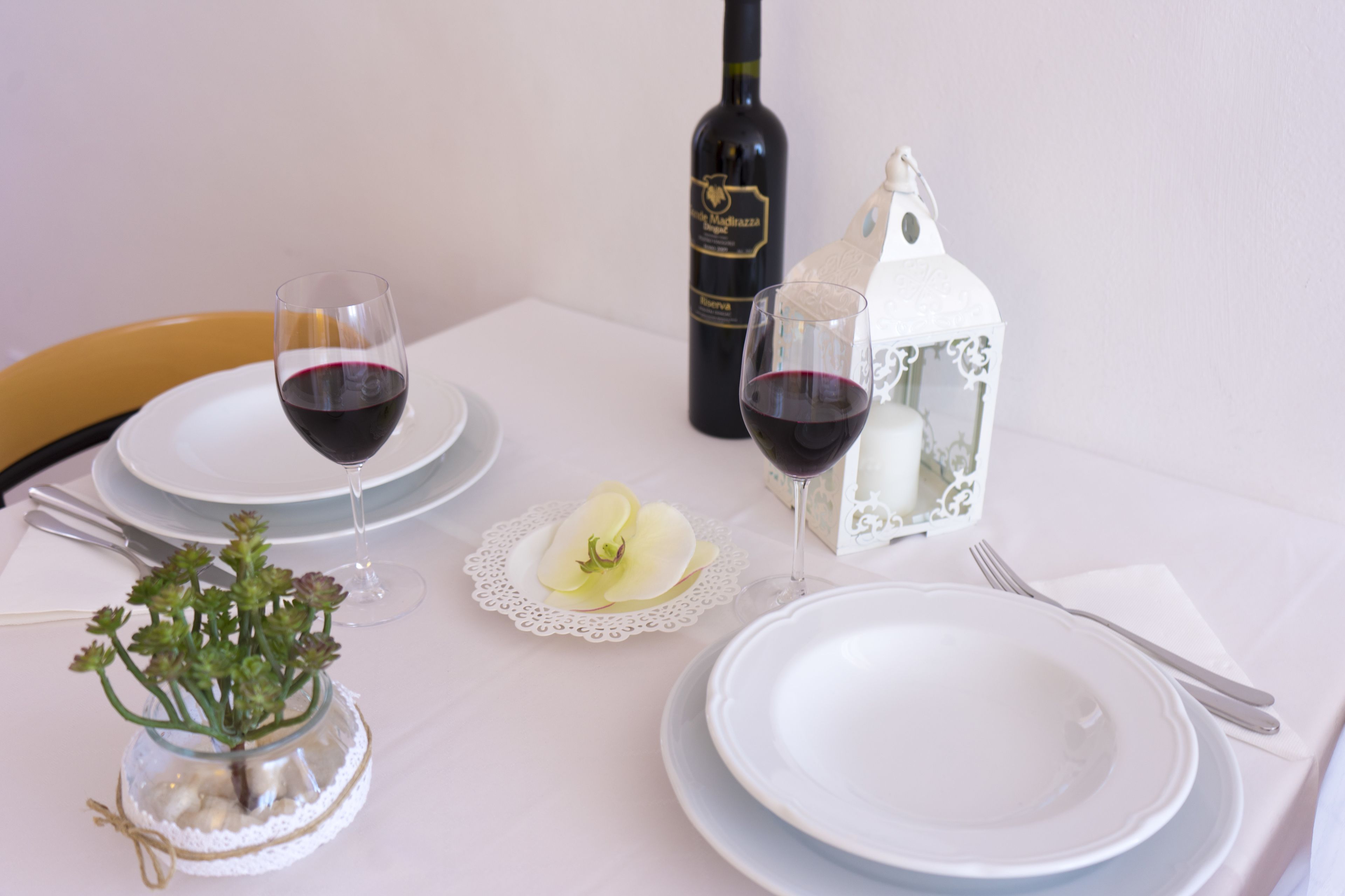 Una mesa dispuesta con platos blancos, dos copas de vino, una botella de vino, un pequeño florero con flores y un farol blanco.