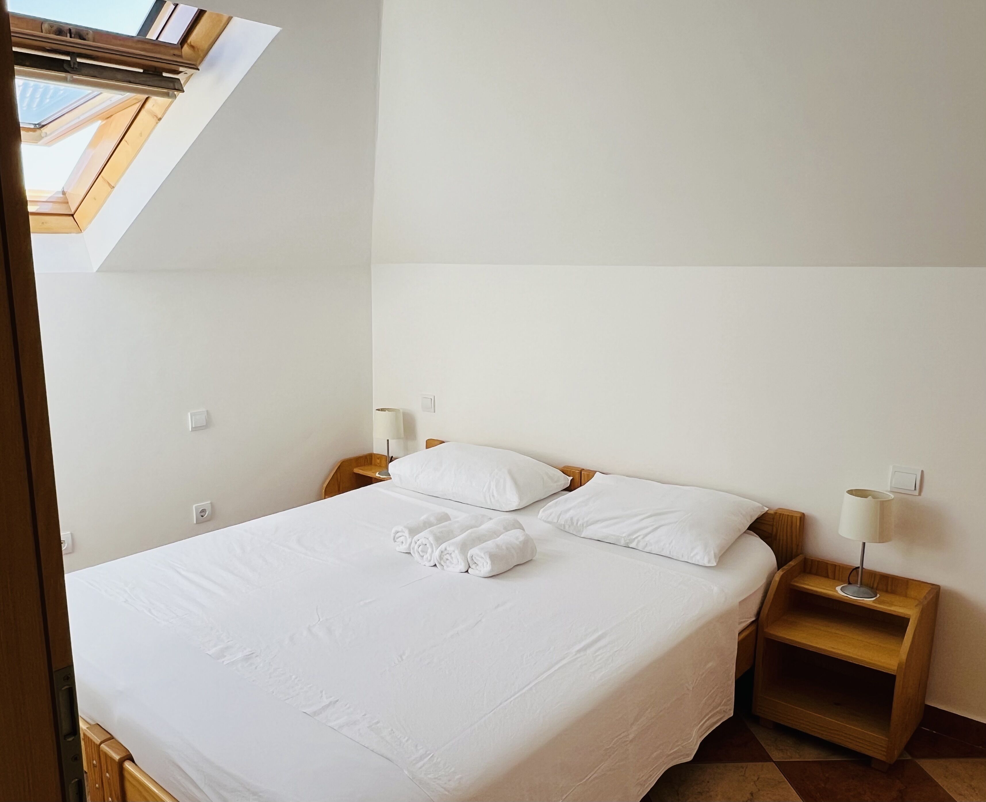 Un dormitorio con tragaluces, una cama con ropa blanca, dos mesitas de noche con lámparas y un cabecero de madera.