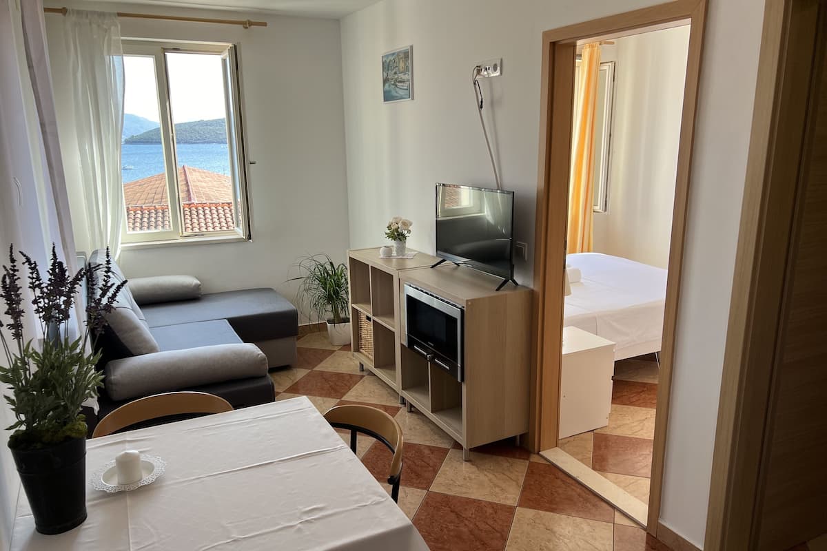 Een compacte hotelkamer met een slaapbank, een televisie op een houten standaard, een eettafel met een witte tafelkleed, en een bed.