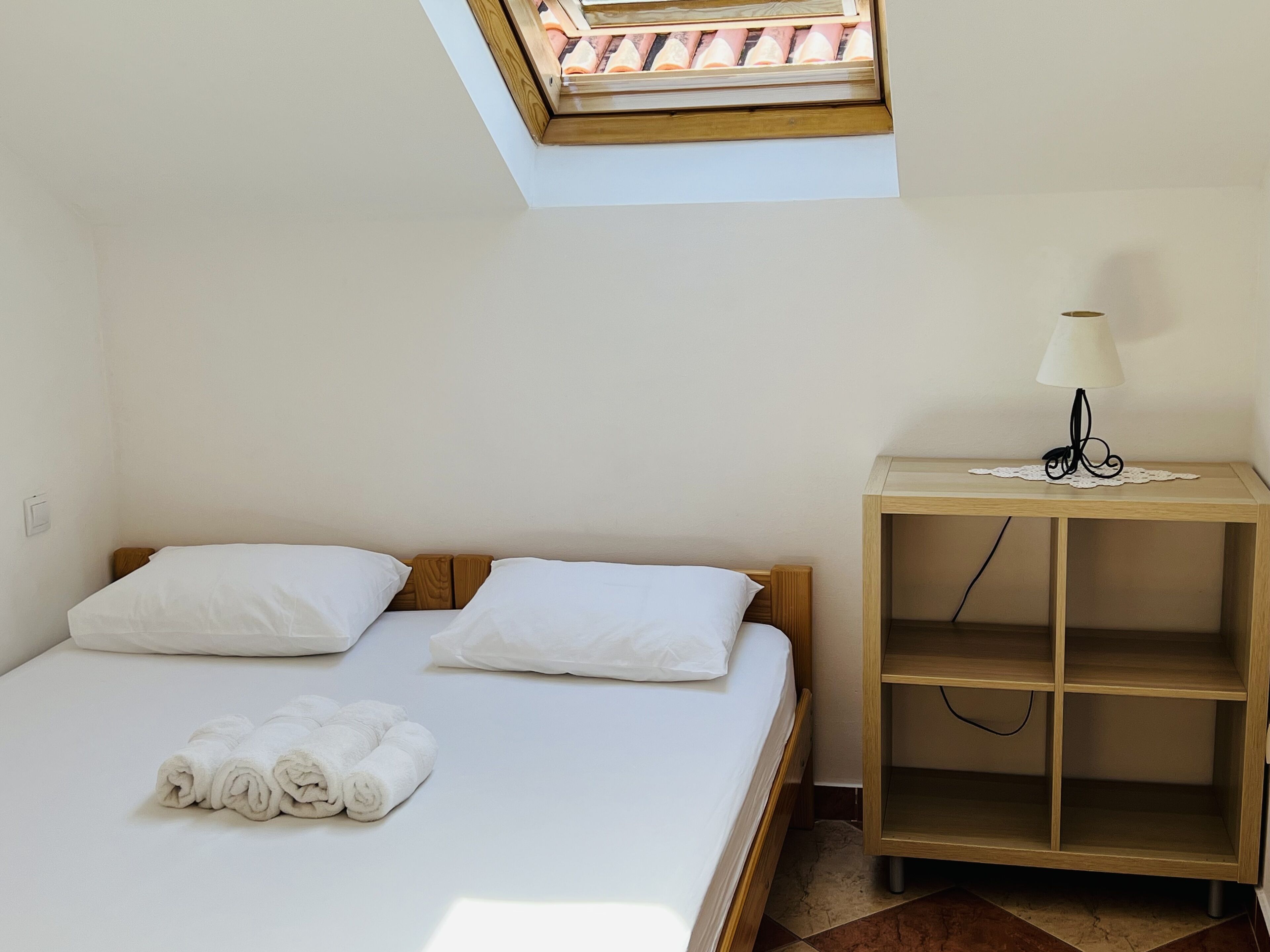 Una cama individual con sábanas blancas y dos almohadas, una mesita de noche de madera con una lámpara, y un tragaluz en el techo.