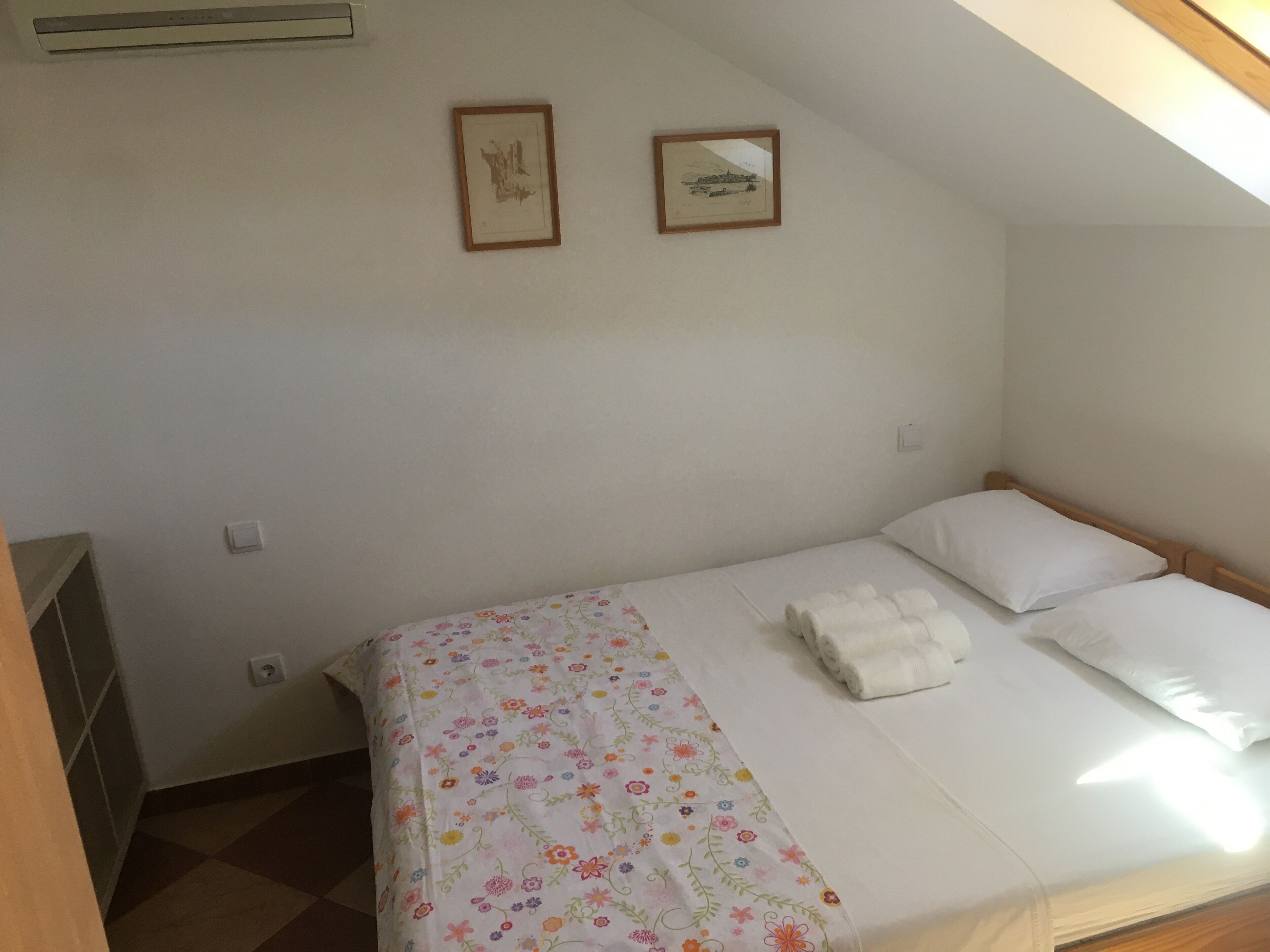 Un lit simple avec une couvre-lit à motifs floraux, deux tableaux encadrés au mur et une fenêtre laissant entrer la lumière naturelle.