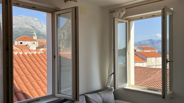 A room with a grey sofa, two open windows, and a view of red-tiled roofs and mountains.