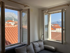 Une pièce avec un canapé gris, deux fenêtres ouvertes et une vue sur des toits couverts de tuiles rouges et des montagnes.