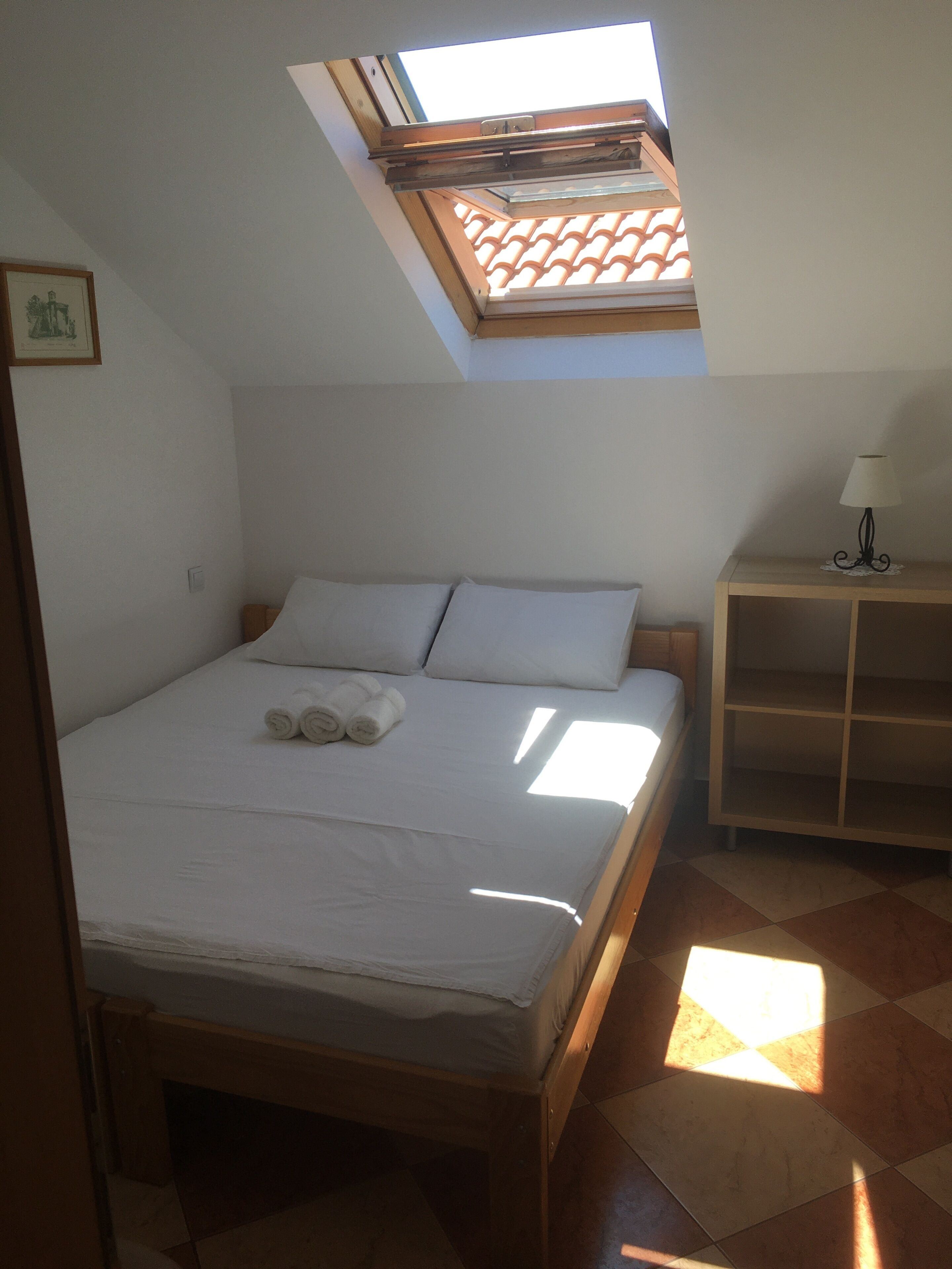 A bedroom with a skylight, a bed with white linens, and a bedside table with a lamp.
