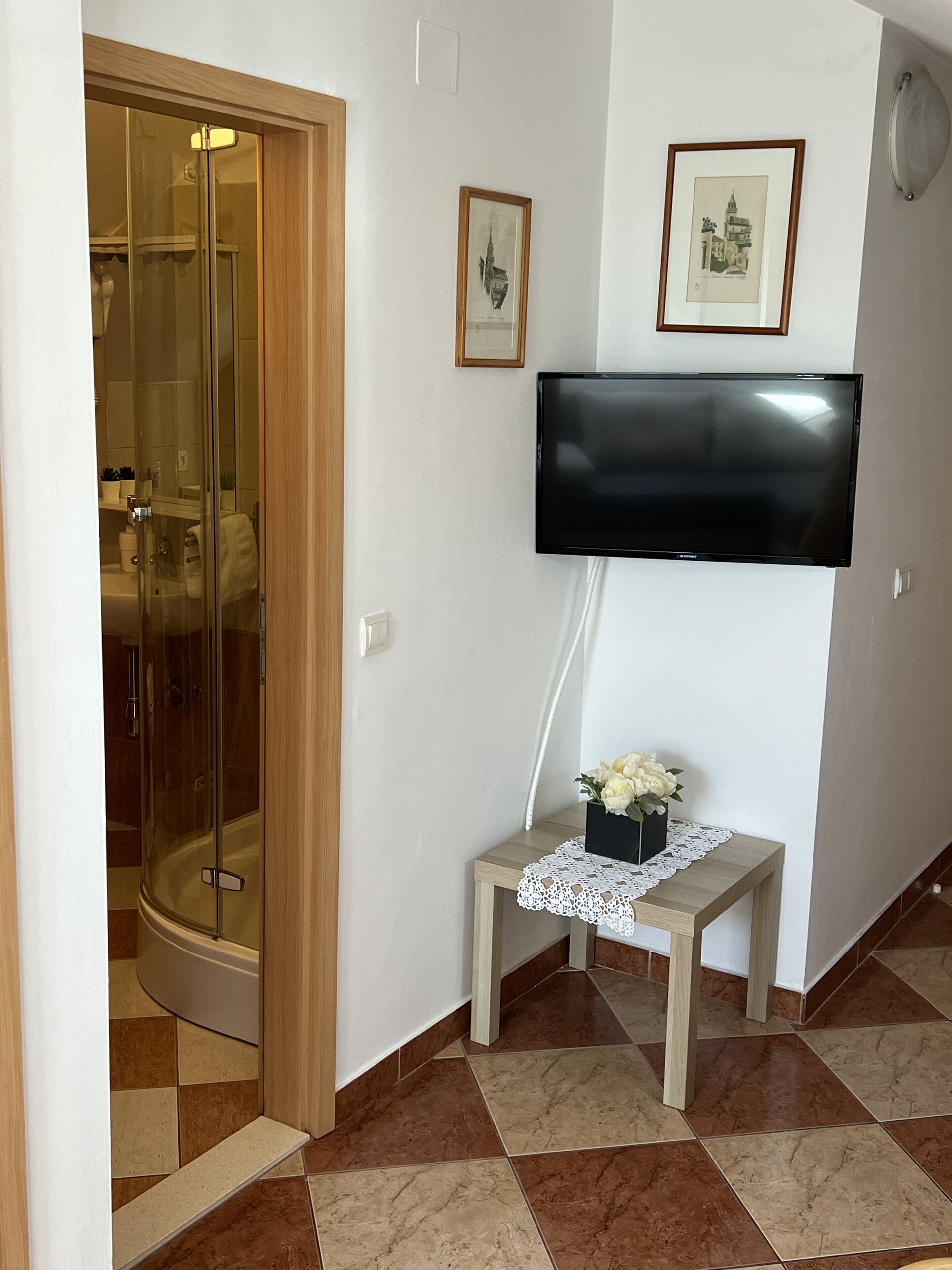 A bathroom with a glass-enclosed shower, a toilet, and a sink. There is a small table with a vase of flowers and a framed picture on the wall.