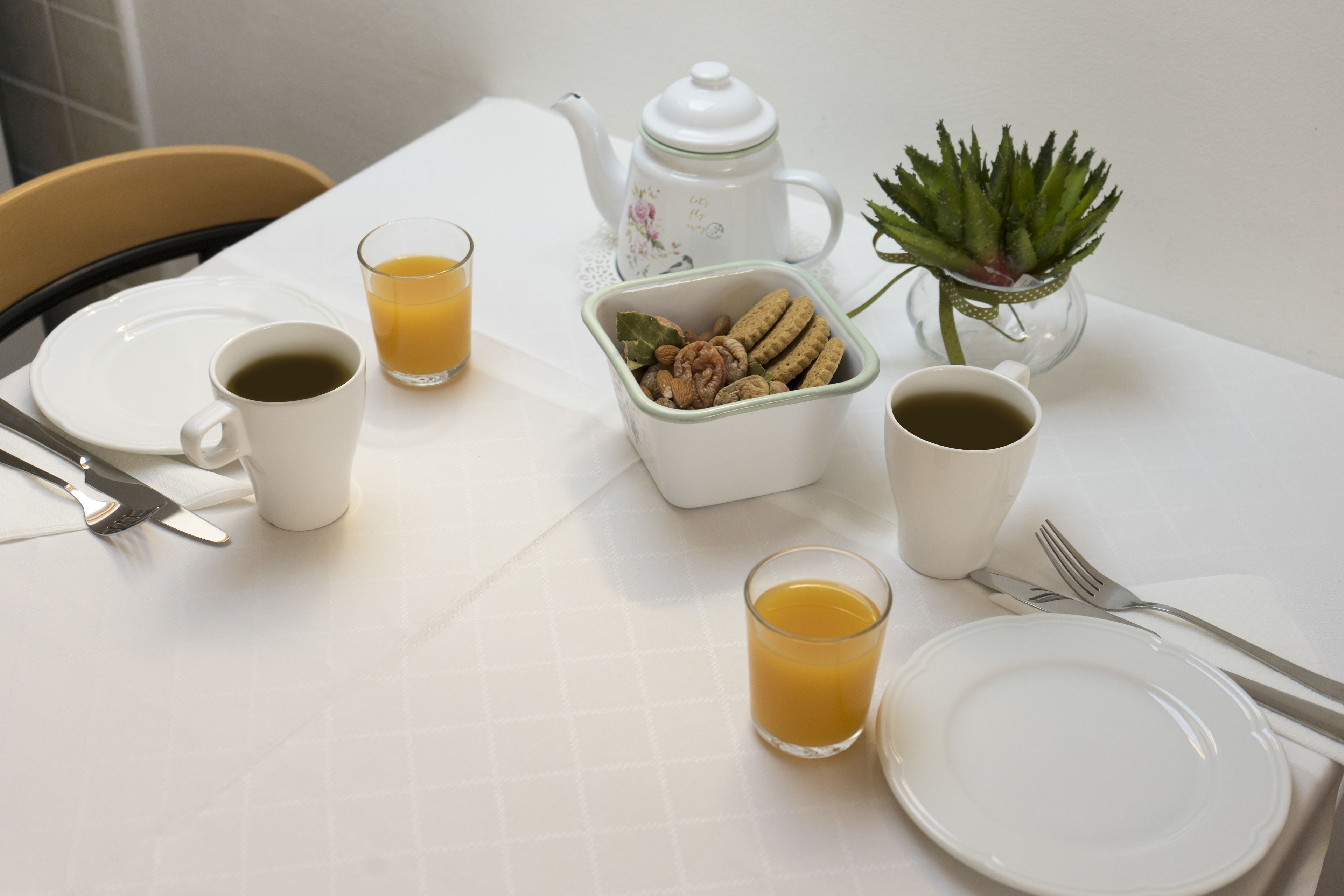 A breakfast table with plates, cups, a teapot, a small plant, and a container of nuts.