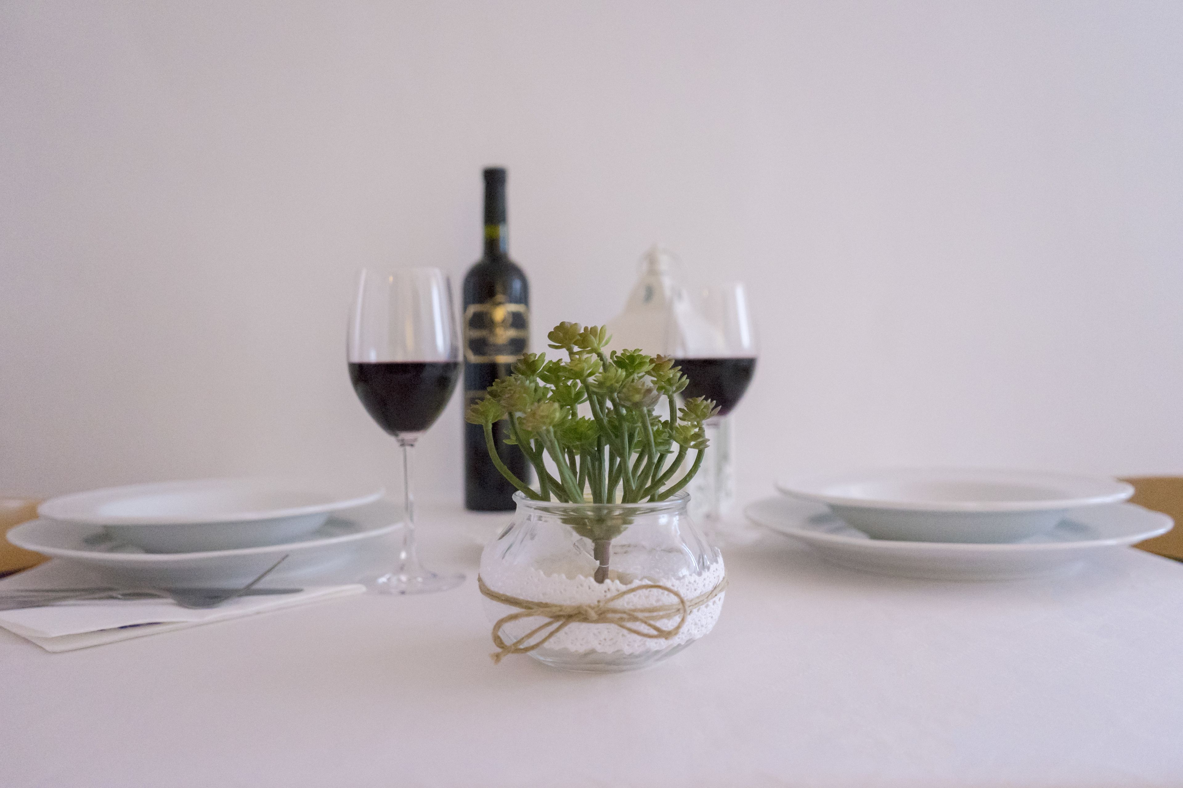 A dining table with a glass vase containing green plants, two wine glasses, and a bottle of wine.