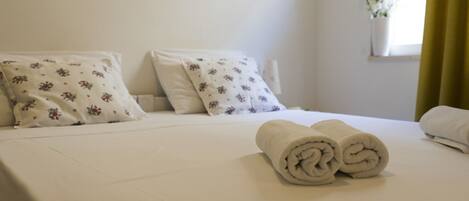 Una cama bien hecha con almohadas estampadas con motivos florales, una toalla enrollada y un jarrón blanco con flores sobre el alféizar de una ventana.