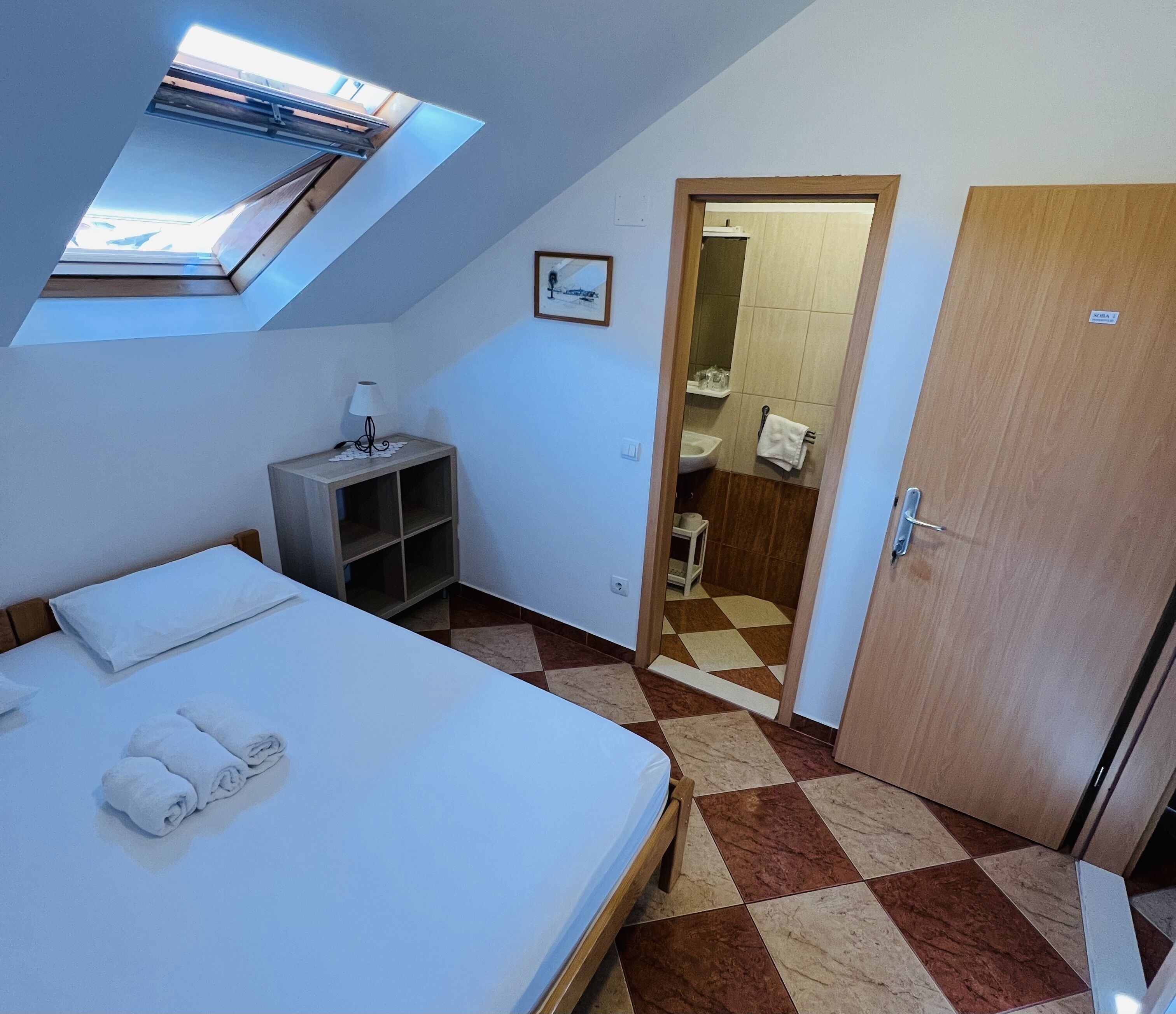 Un dormitorio con tragaluz, una cama con toallas, una mesita de noche, un mueble pequeño y un baño visible a través de una puerta abierta.