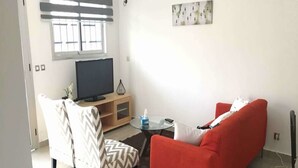 A living room with a red sofa, a white armchair, a TV on a wooden stand, and a small table with a glass top.
