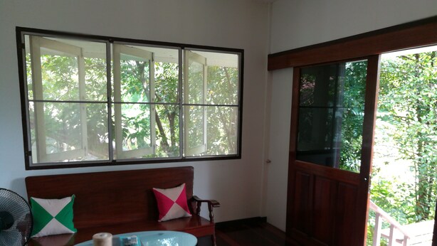 A room with a wooden floor, a glass table with a candle, a wooden bench with cushions, and a large window with a view of trees.