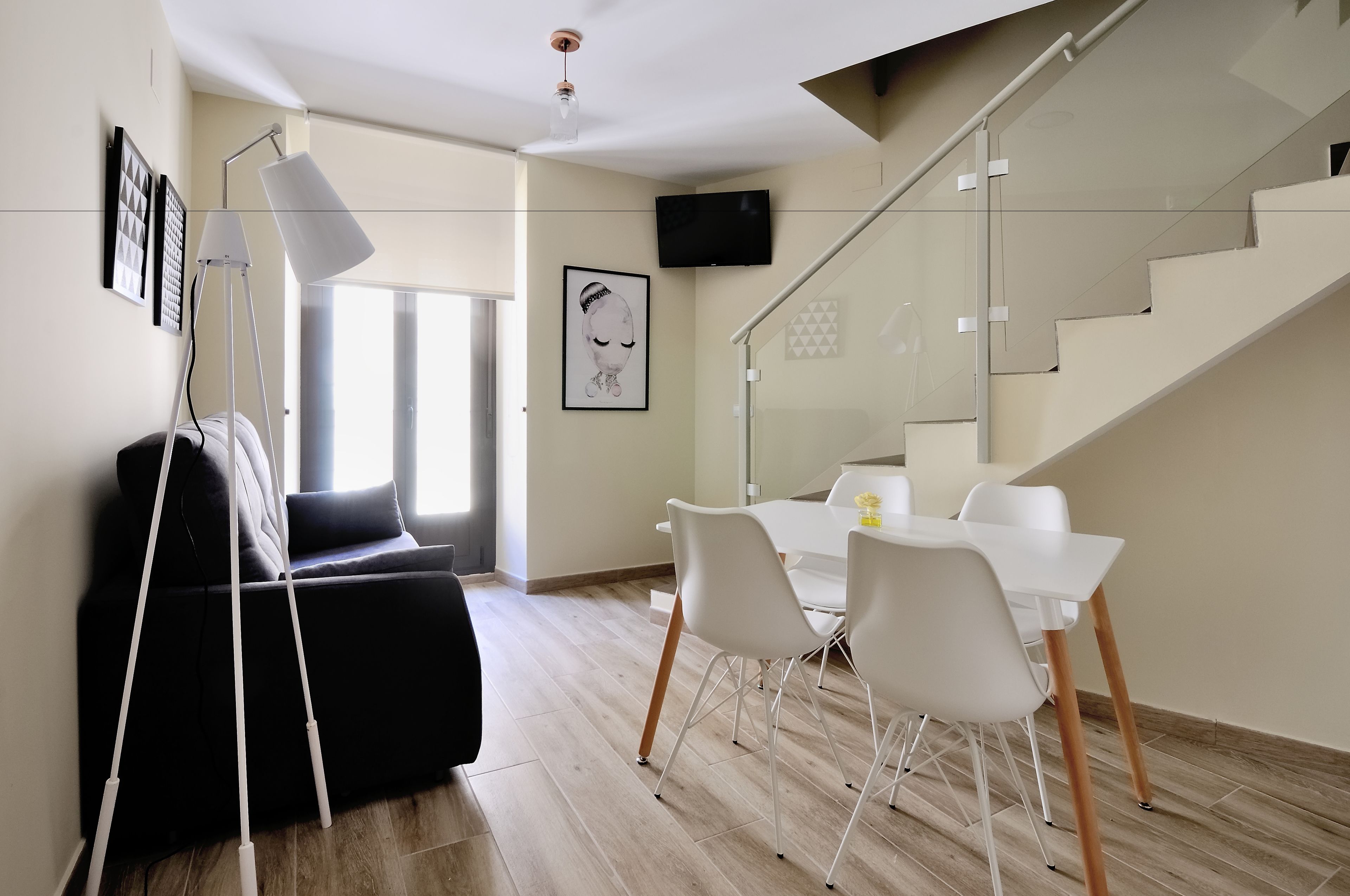 Una sala moderna con un sofá negro, una escalera de cristal y un comedor con sillas blancas y una mesa.