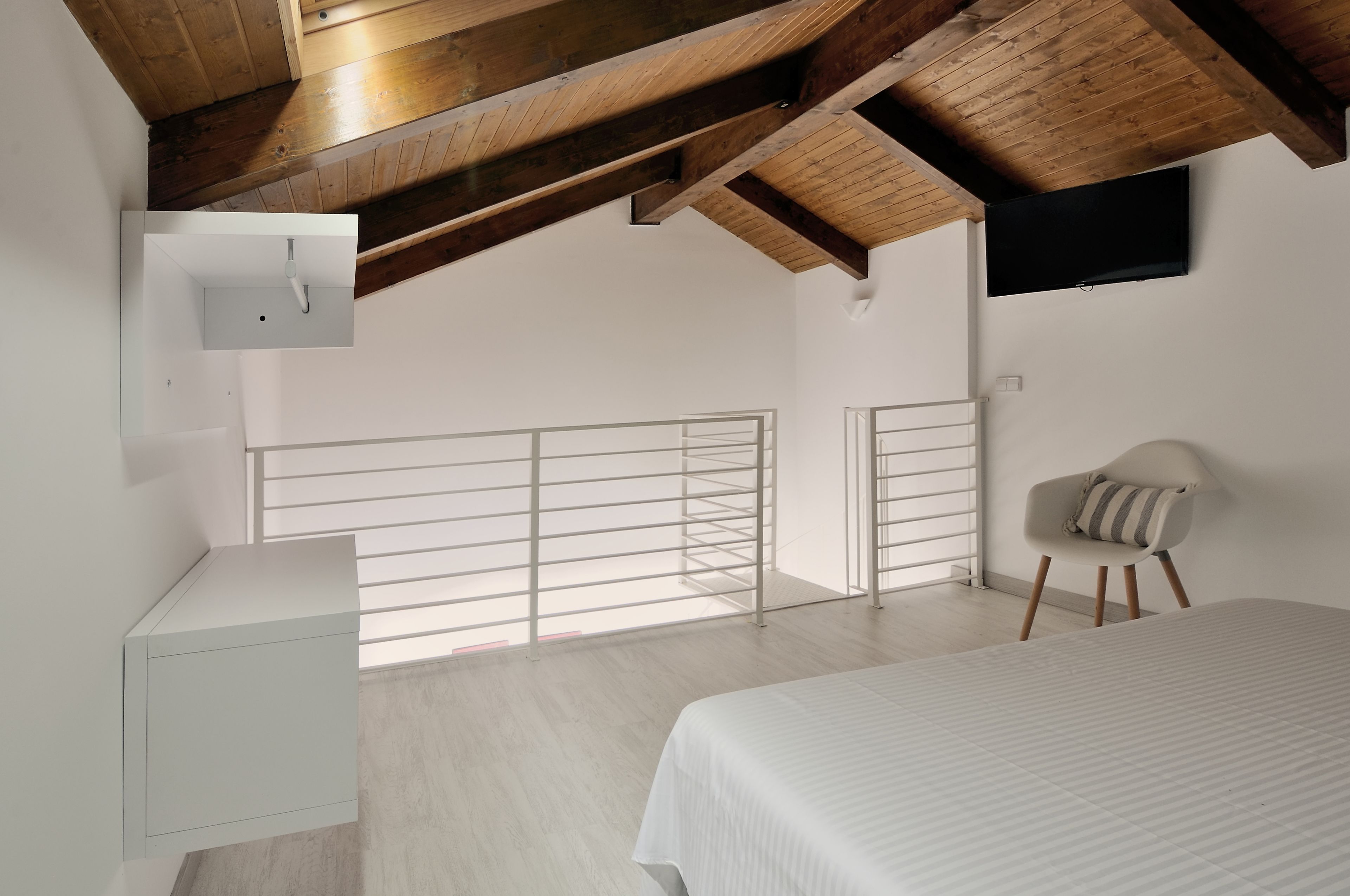 Une chambre avec un lit, une petite table de chevet, une chaise, un téléviseur fixé au mur et un balcon avec une balustrade.