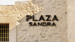 Ein Schild mit der Aufschrift 'PLAZA SANDRA' an einer strukturierten Wand, vor der sich ein gefliester Pool befindet.