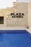 Uma placa com a inscrição 'PLAZA SANDRA' em uma parede texturizada, com uma piscina azulejada à frente.
